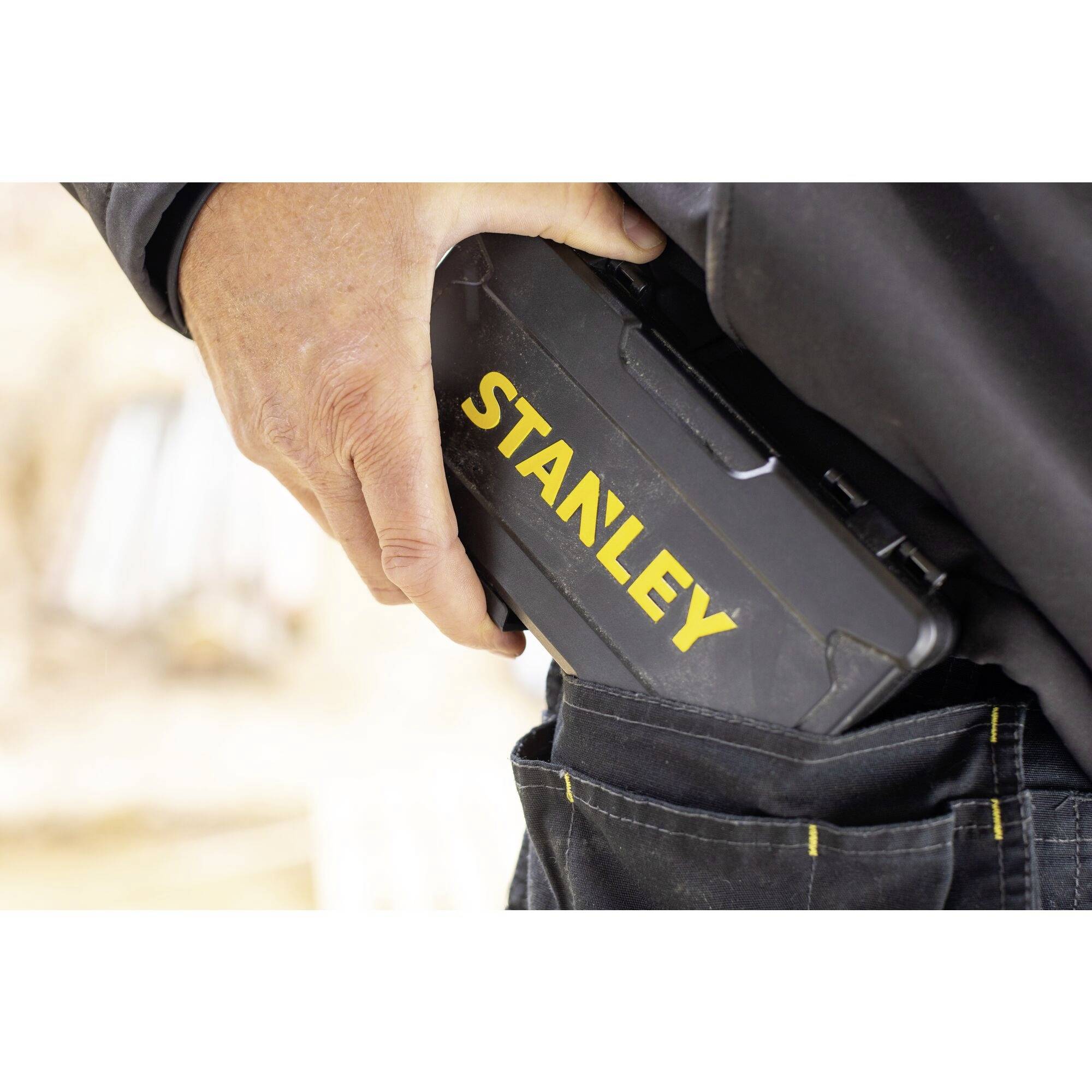 Een persoon stopt een zwarte, rechthoekige Stanley-doos in de broekzak. De doos is voorzien van gele tekst.