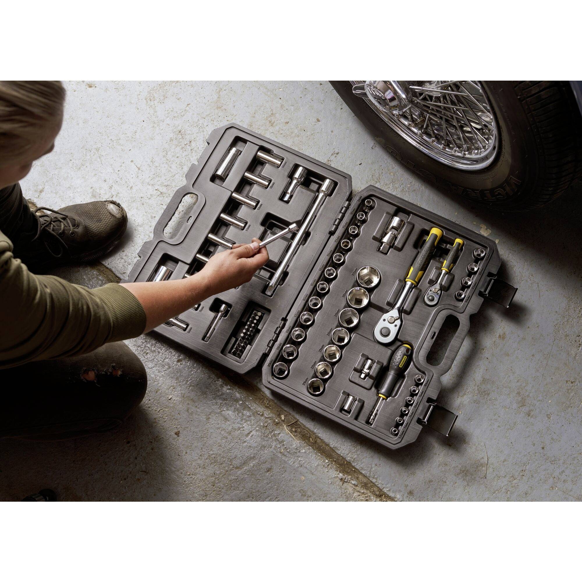 Een persoon controleert een geopende gereedschapskist met verschillende dopsleutels en gereedschappen op een betonnen vloer, naast de band van een voertuig.