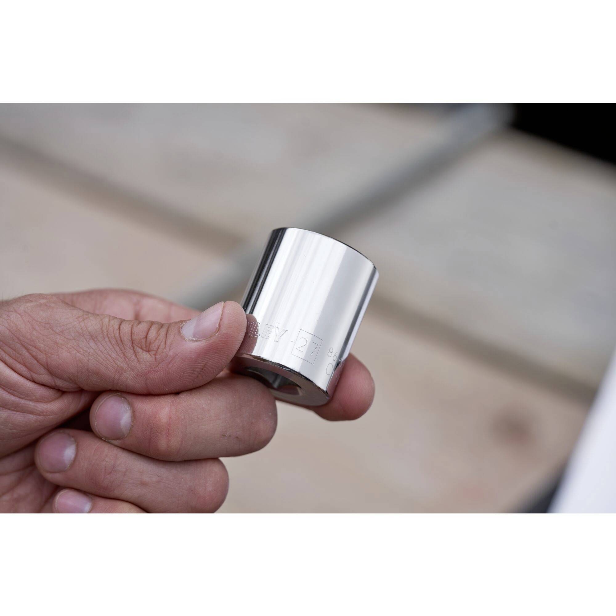 Een hand houdt een zilveren, metalen opzetstuk vast, mogelijk een gereedschap. De achtergrond is onscherp en toont een werkoppervlak.