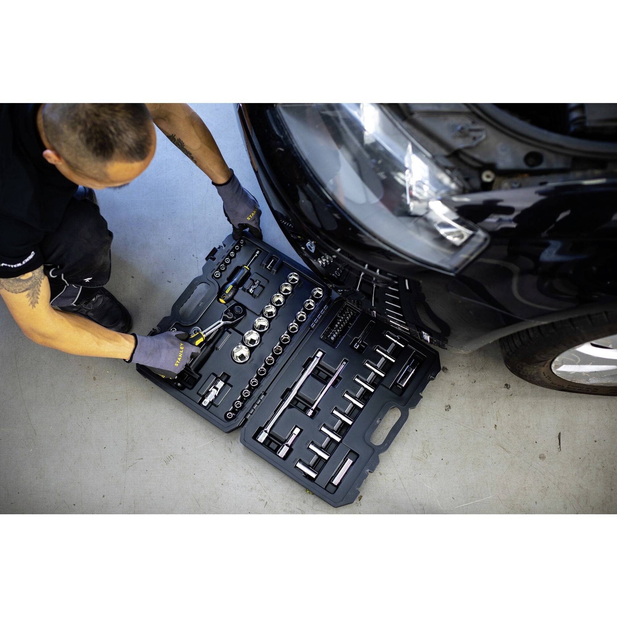 Een monteur in zwarte kleding werkt aan een auto naast een open gereedschapskist met verschillende moersleutels.