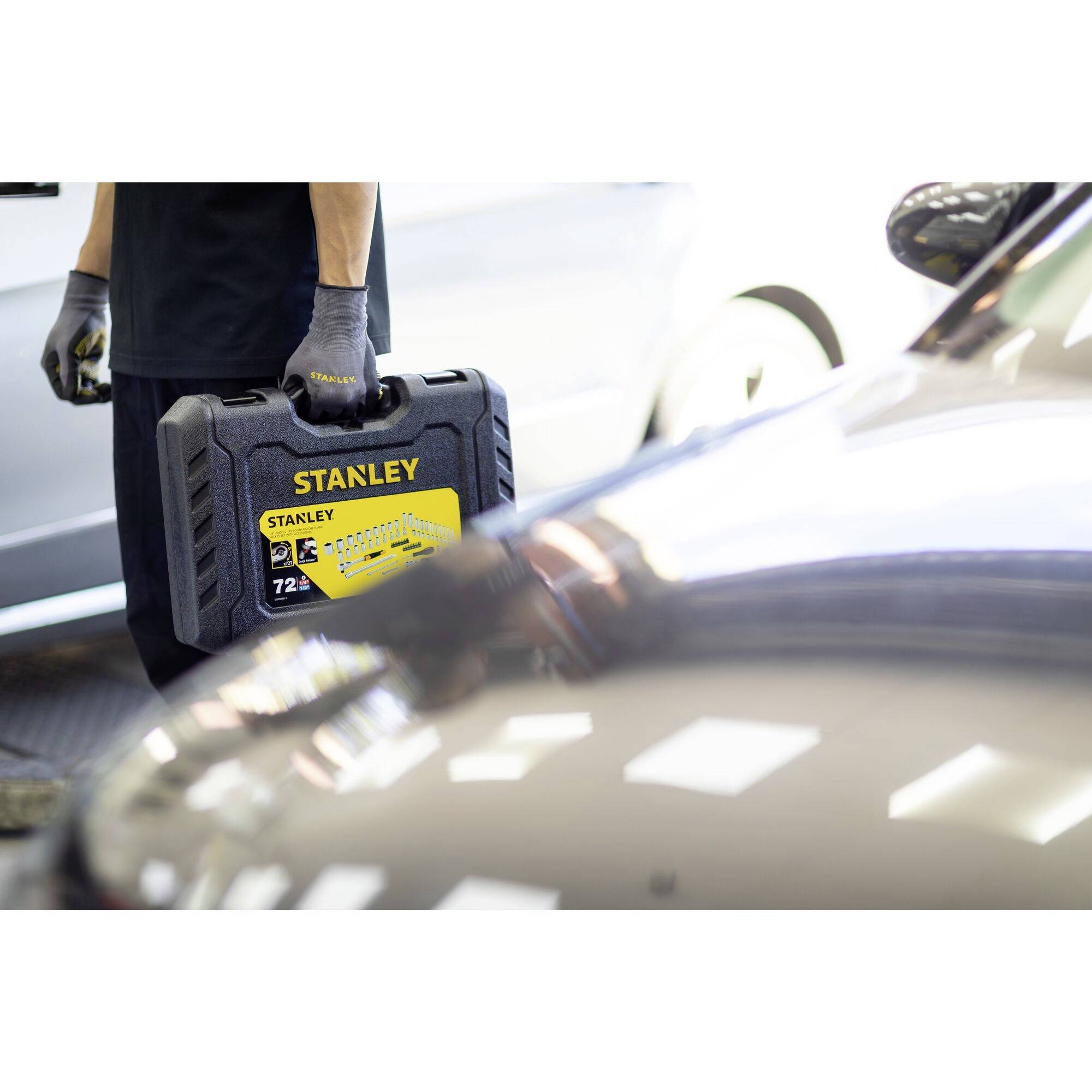Een persoon houdt een Stanley-gereedschapskist naast een auto. Lichtreflecties zijn zichtbaar op het voertuig.