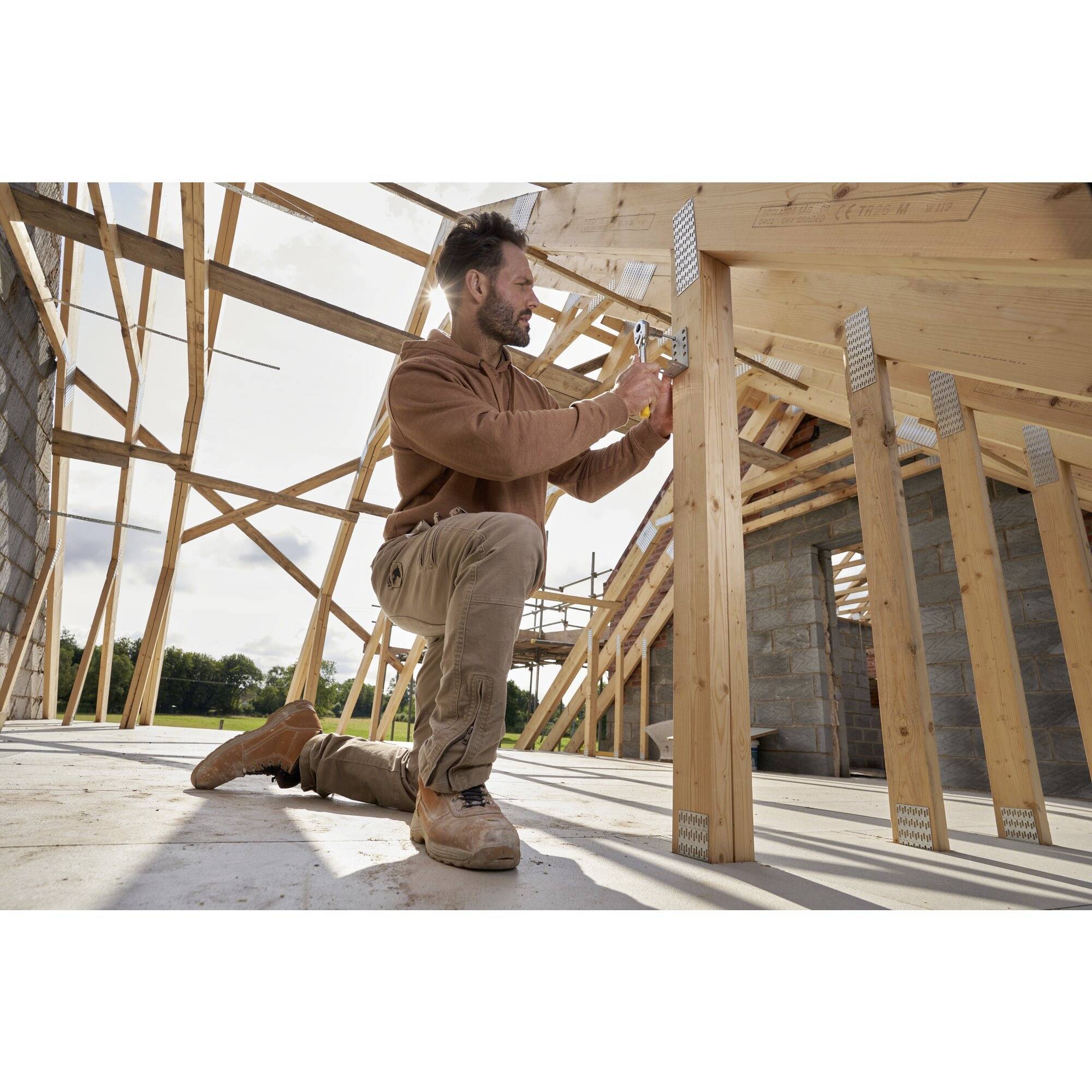 Een man die op een geknield been werkt, bevestigt houten frames in een gedeeltelijk opgebouwd gebouw. Steiger omringt de structuur.