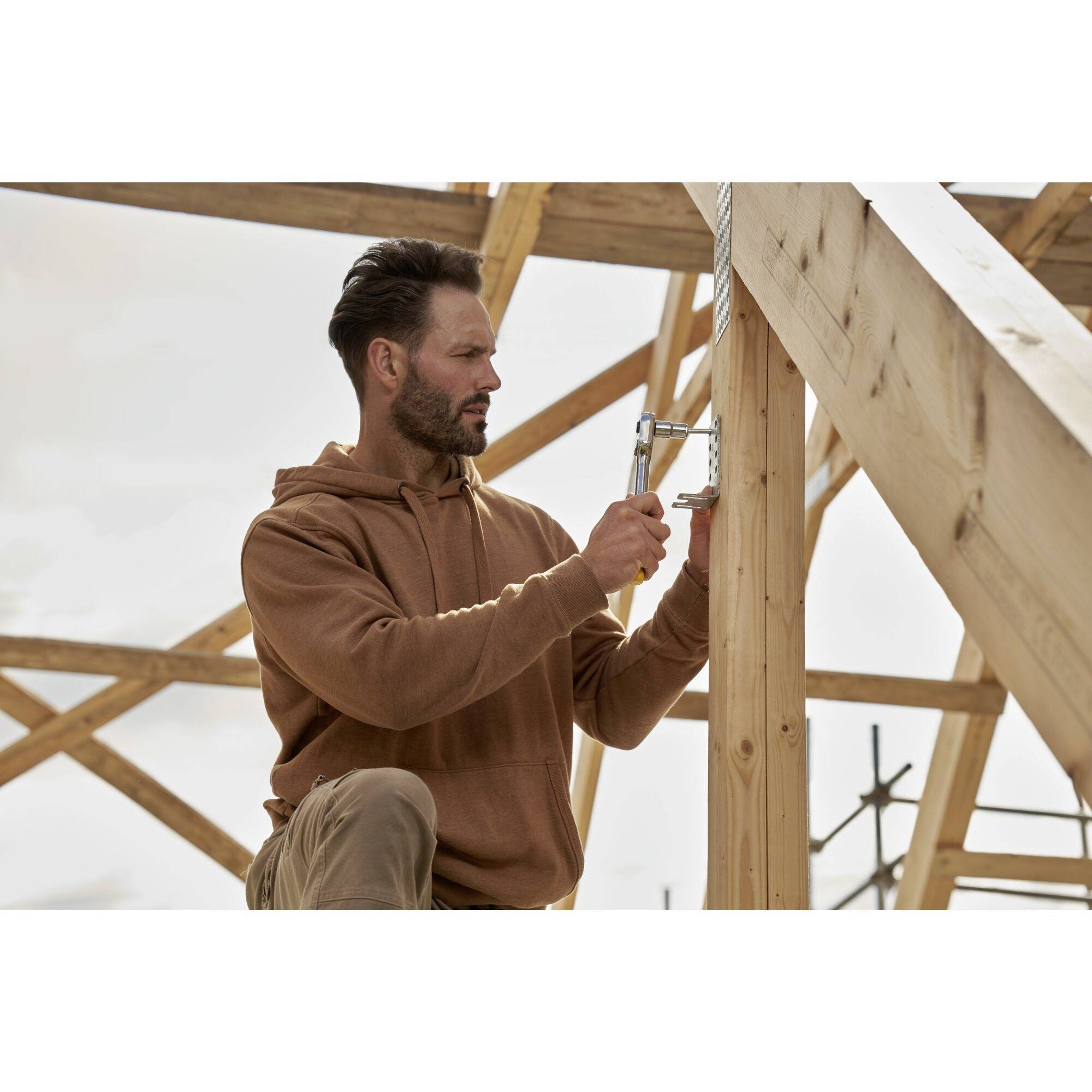 Een man in een bruine hoodie werkt aan een houten constructie op een bouwterrein. Hij gebruikt een gereedschap om een touw te bevestigen.