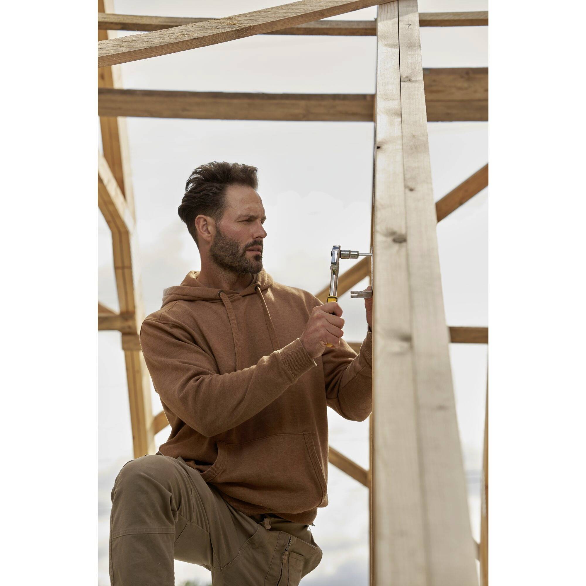 Een man hamert een spijker in een houten balk van een onvoltooid houten frame. Hij draagt een bruine hoodie.