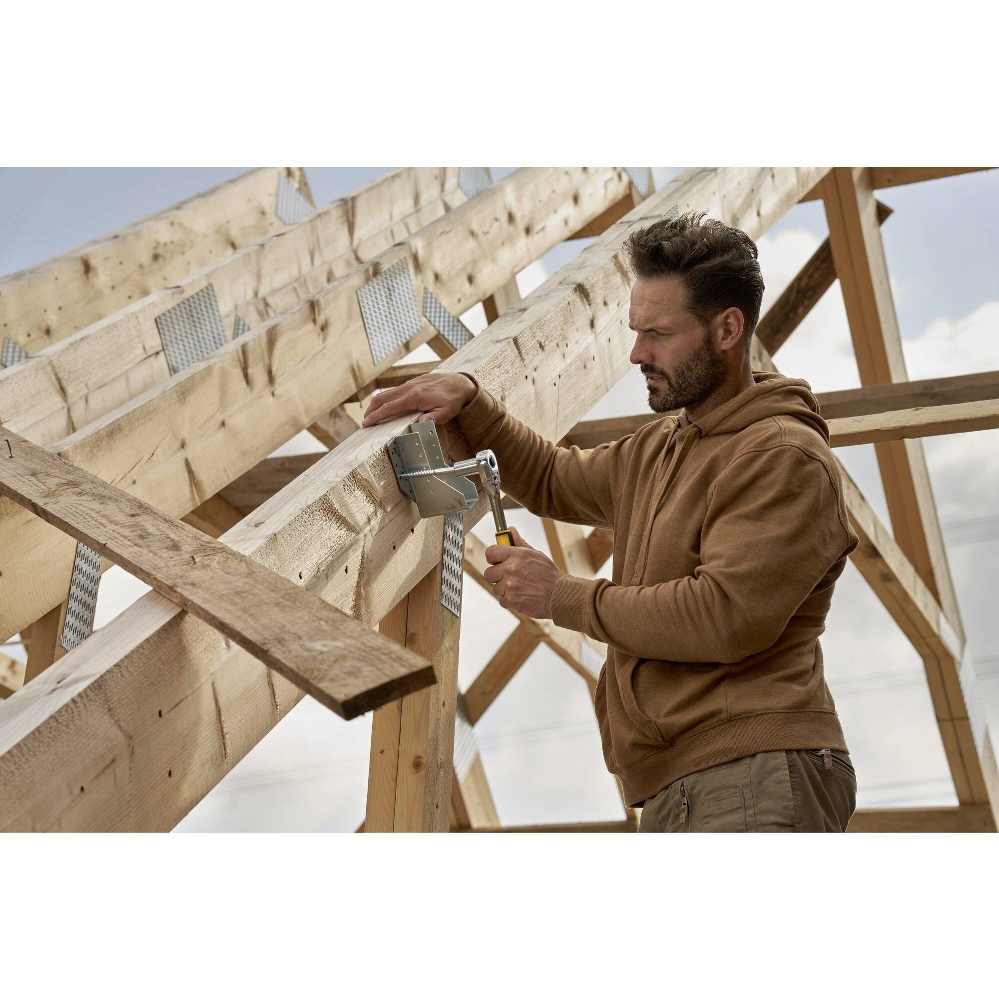 Een man in een bruine hoodie werkt met een gereedschap aan een houten frame van een gebouw in de open lucht.