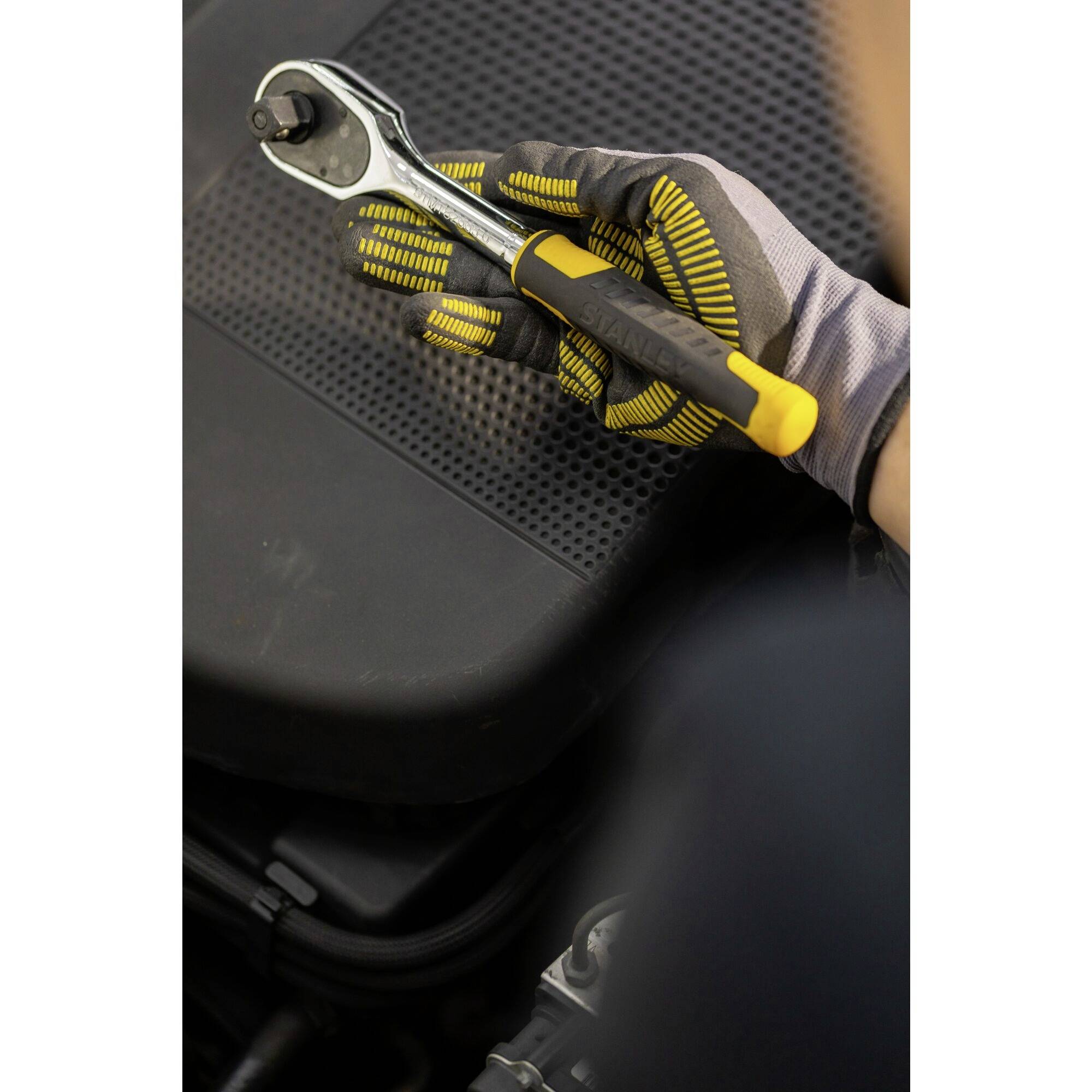 Een persoon met werkhandschoenen houdt een ratel-steeksleutel boven een motorruimte, klaar voor reparatie.