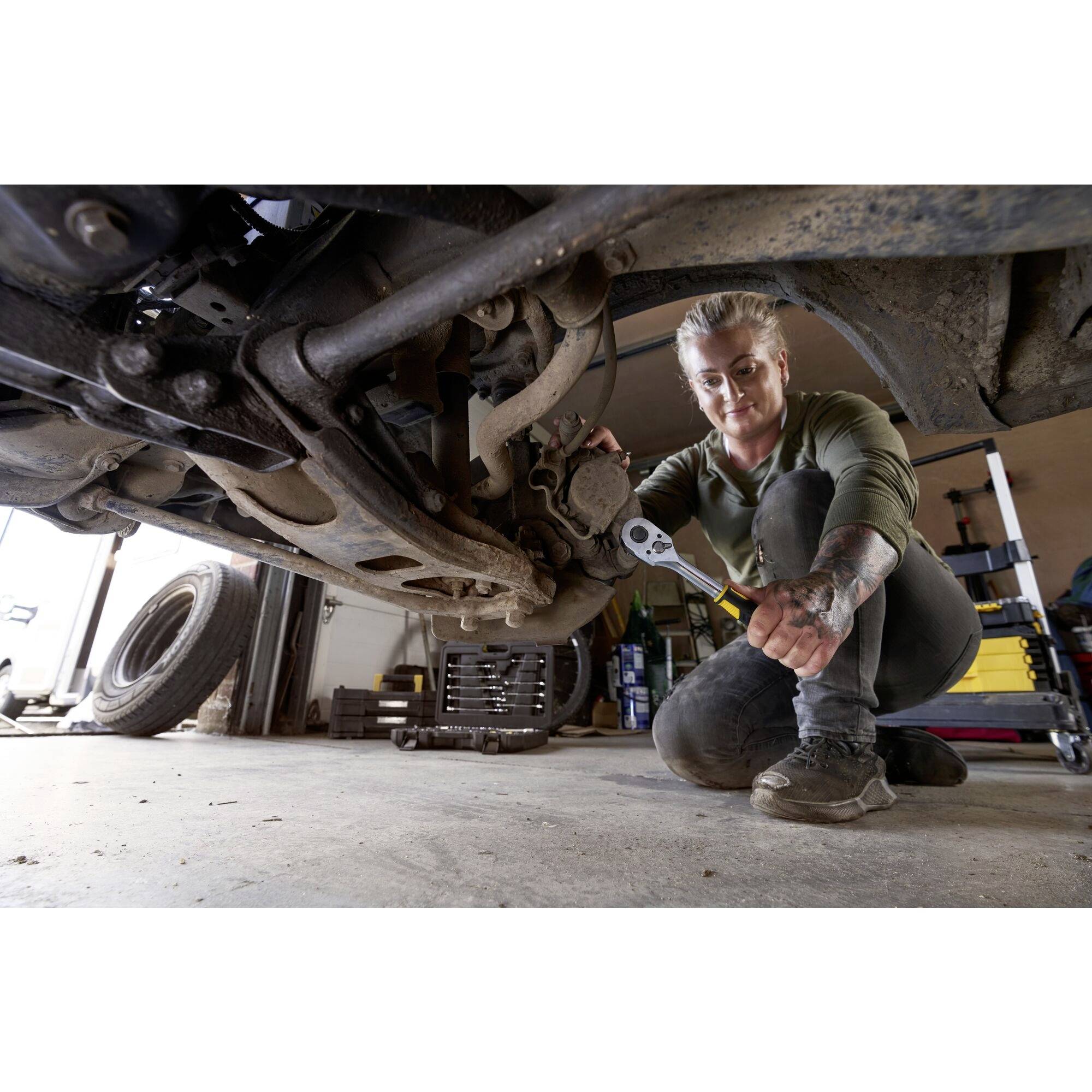 Een monteur werkt met gereedschap onder een voertuig in een garage, toont een reparatie- of onderhoudssituatie.