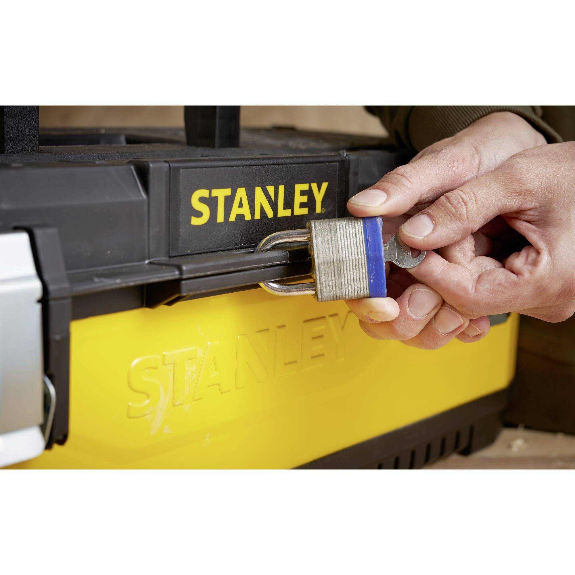 Twee handen sluiten een hangslot op een zwarte Stanley-gereedschapskist. Het slot beveiligt de kist.