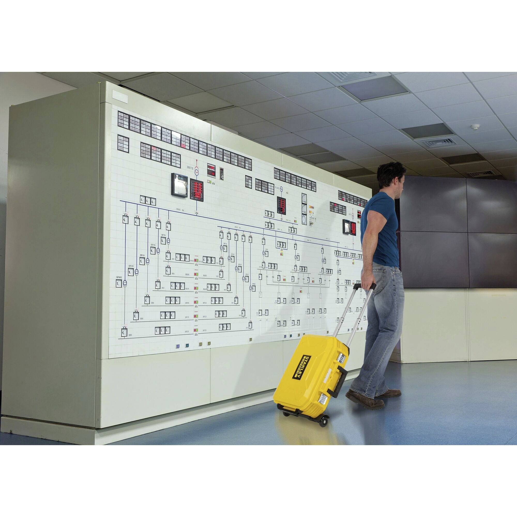 Een persoon trekt een gele koffer voor een groot controlepaneel in een technische controlekamer.