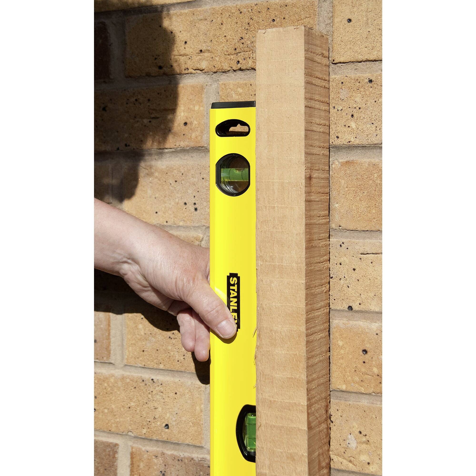 Een hand houdt een gele waterpas en een houten plank tegen een bakstenen muur om de verticale uitlijning te controleren.