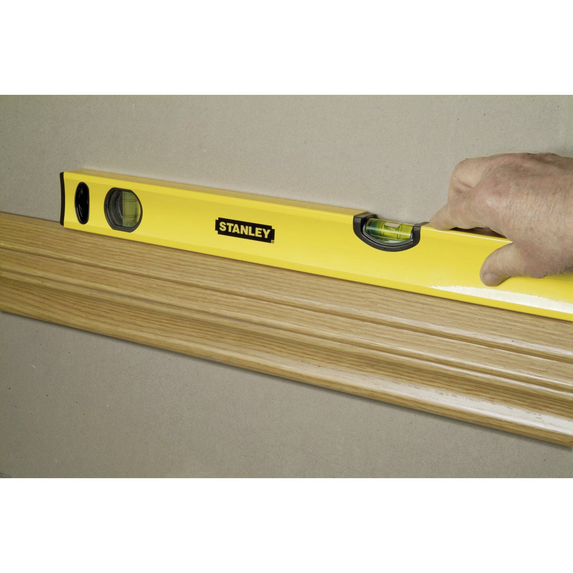 Een gele waterpas wordt langs een houten plank gelegd. Een hand houdt de waterpas vast.