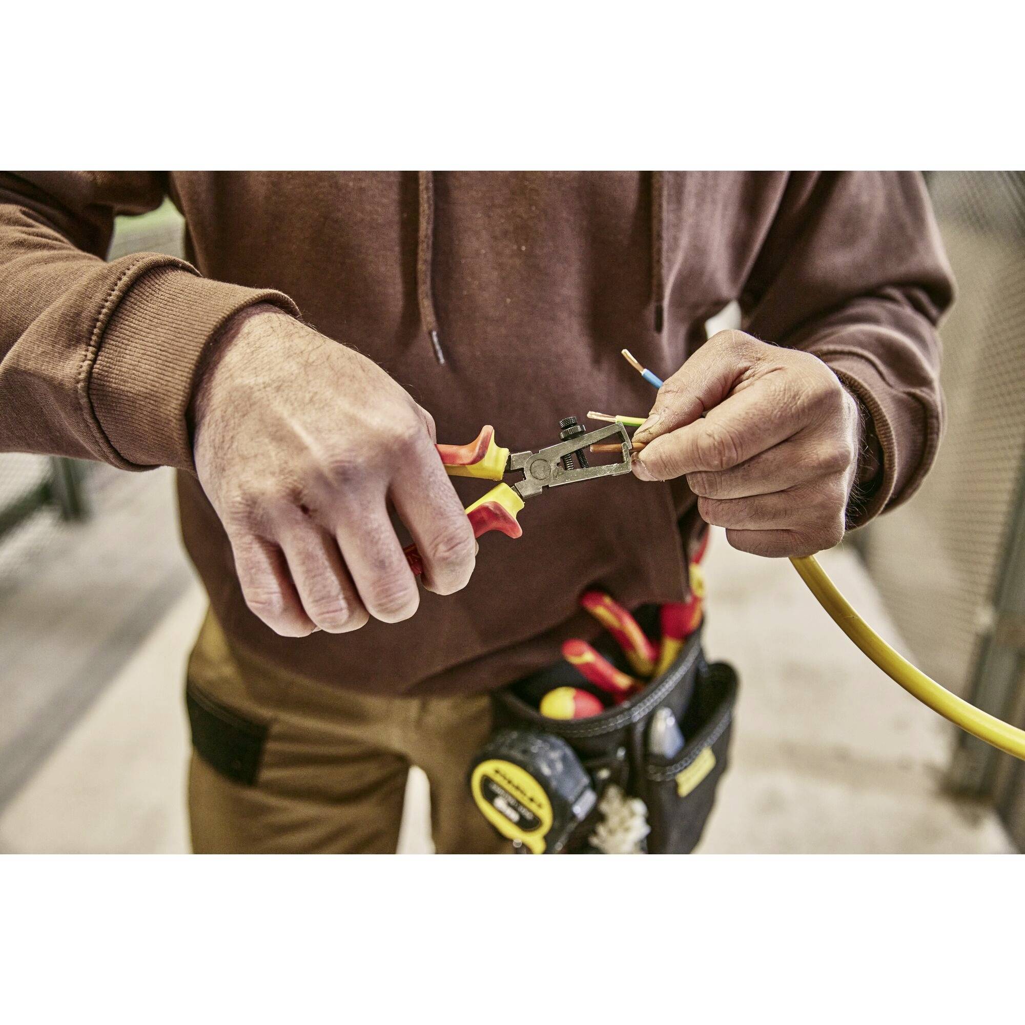Een persoon knipt een geel elektrisch kabel door met een gereedschap. Ze draagt een bruine trui en heeft een gereedschapsriem.