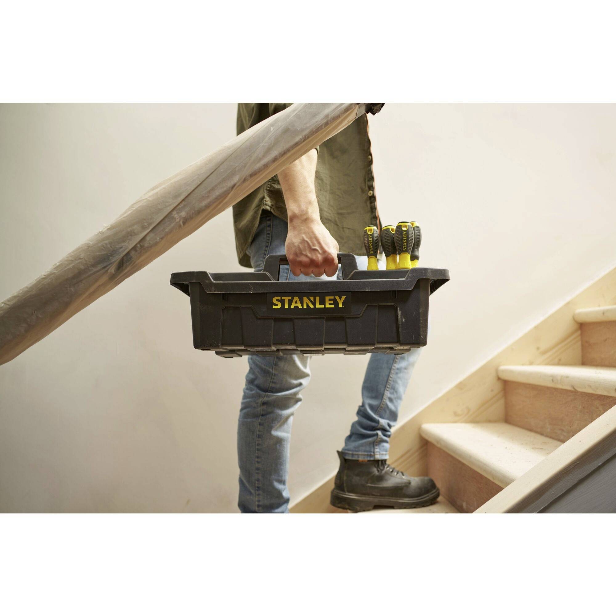 Een persoon draagt een 'Stanley'-gereedschapskist een trap op. De scène toont doe-het-zelfwerkzaamheden in een woonomgeving.