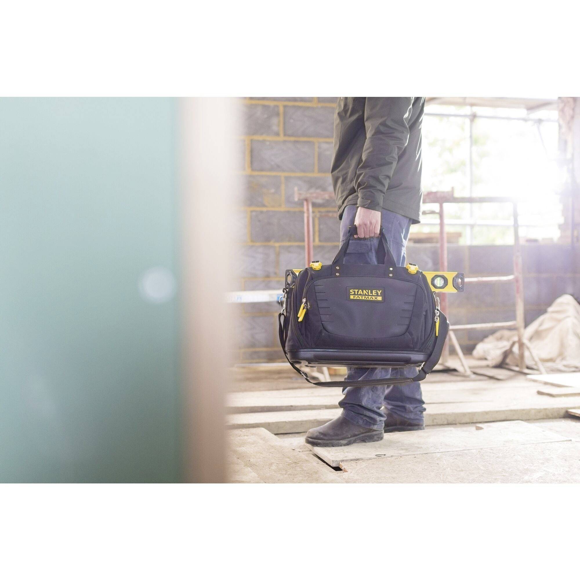 Een persoon draagt een gereedschapstas in een onafgewerkte ruimte met bakstenen muren.