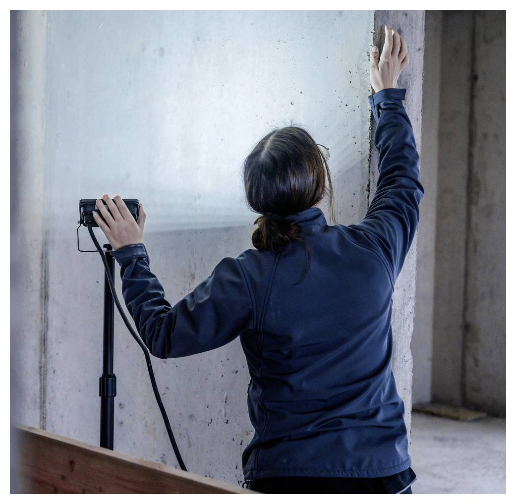 Een persoon in een donkere jas meet of markeert een betonnen muur met een handheld apparaat, staande binnenshuis in de buurt van onafgewerkte constructieoppervlakken.