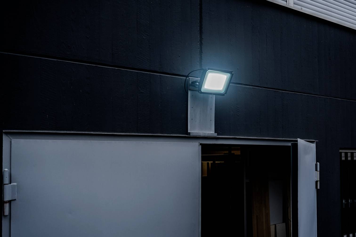 Een moderne buitenlamp schijnt boven een gedeeltelijk geopende deur aan een donker gebouw en verlicht de ingang.