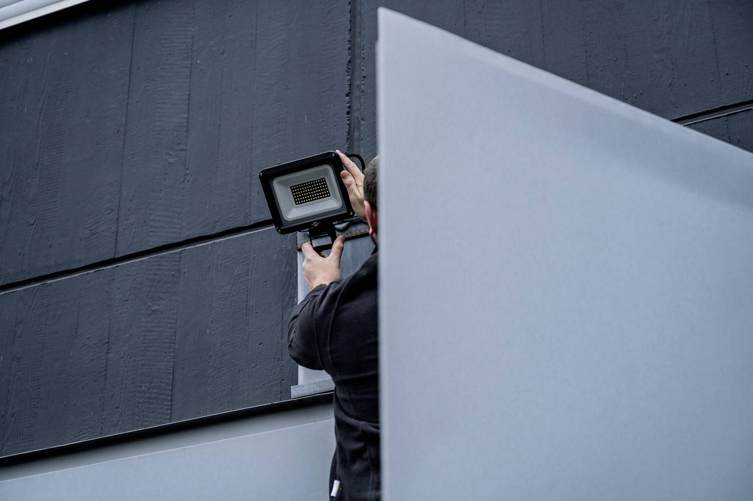 Een persoon installeert een LED-schijnwerper aan een donkere muur. De scène toont technisch werk op het gebied van verlichtingstechniek.