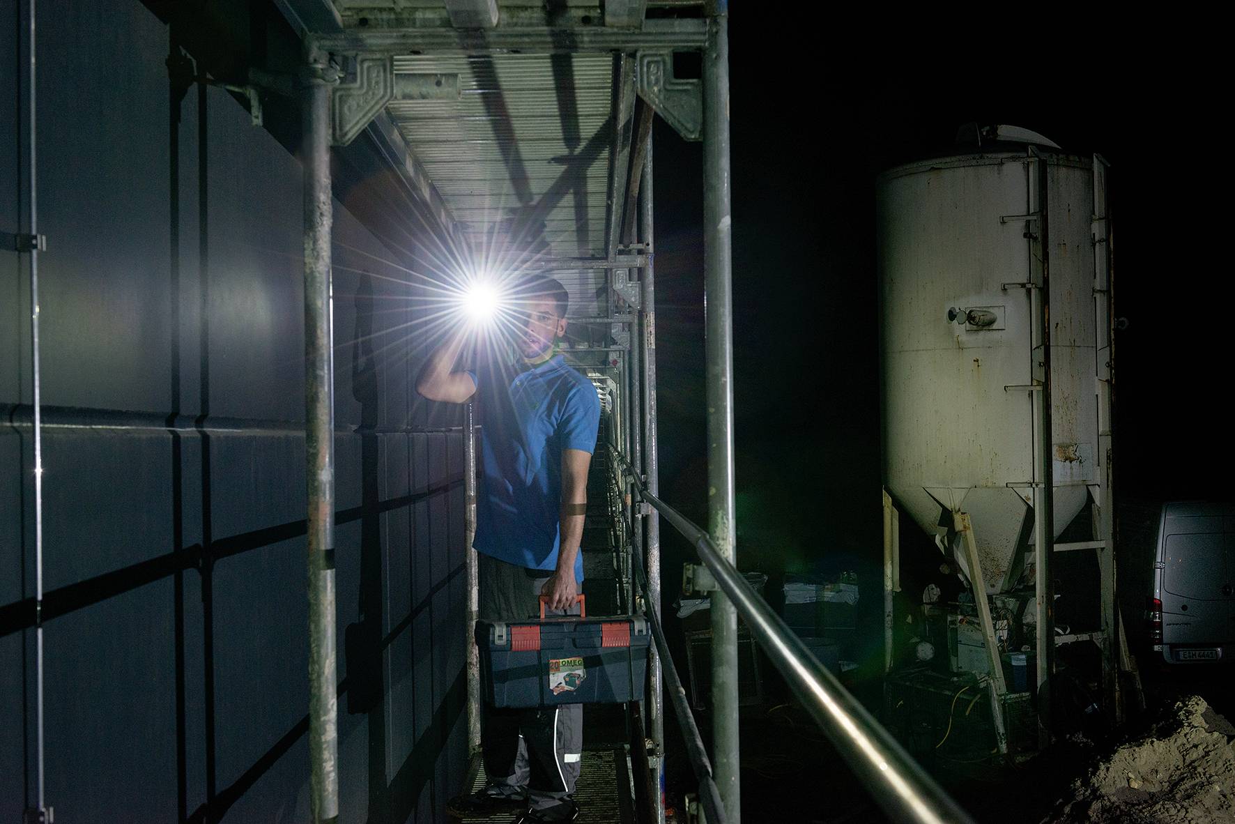 Een arbeider staat 's nachts op een steiger en schijnt met een zaklamp. Hij draagt een gereedschapskist. Op de achtergrond zijn industriële installaties zichtbaar.