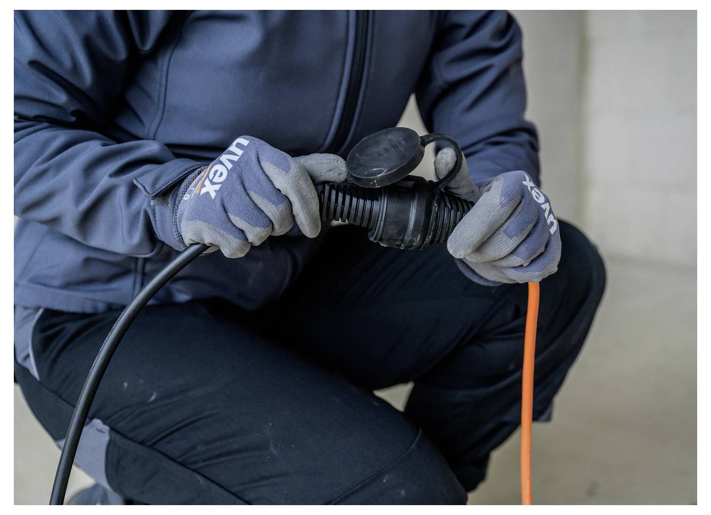 Persoon met handschoenen die een zwarte kabel aansluit op een ronde stekker of adapter, met een zichtbare oranje kabel, zittend binnenshuis.