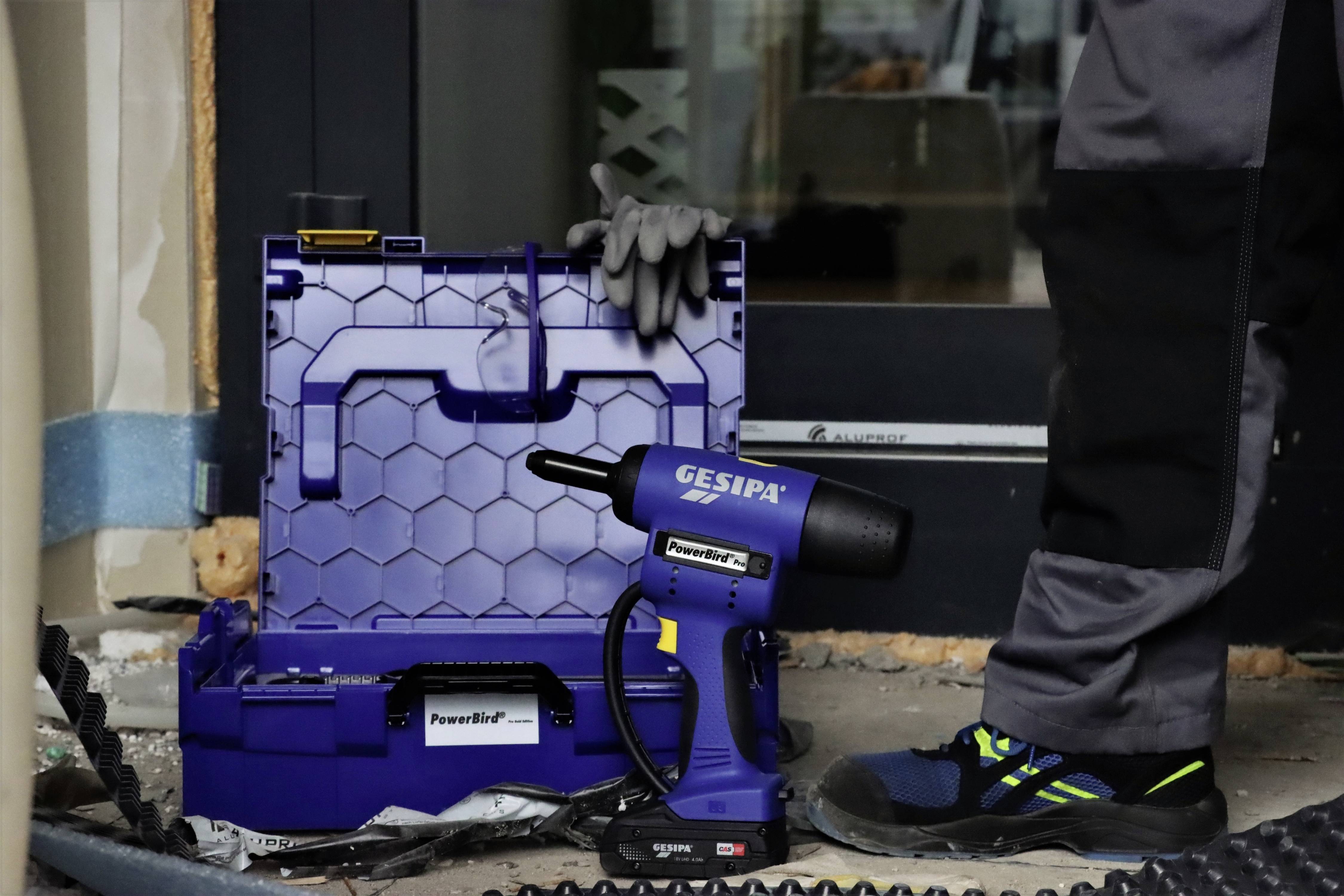 Een persoon staat naast een geopende gereedschapskist met een elektrisch popnagelpistool. Op de achtergrond is een gebouw zichtbaar.