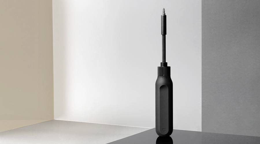 Een zwarte schroevendraaier met een lange steel staat op een reflecterende oppervlakte, voor een achtergrond van grijs- en beige tinten.