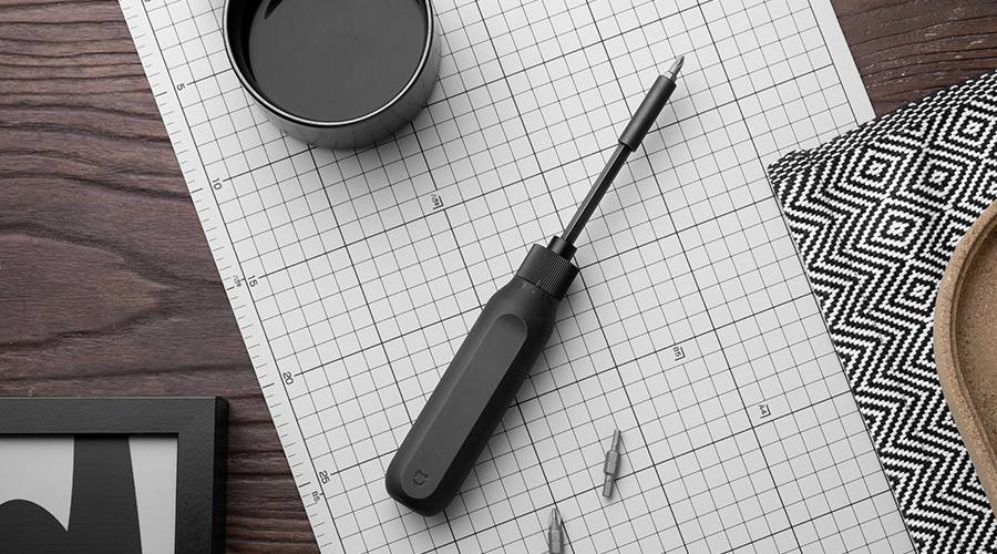 Een schroevendraaier ligt op geruit papier, omringd door schroevendraaierbitjes. Links ernaast staat een beker en rechts een stofmonster.