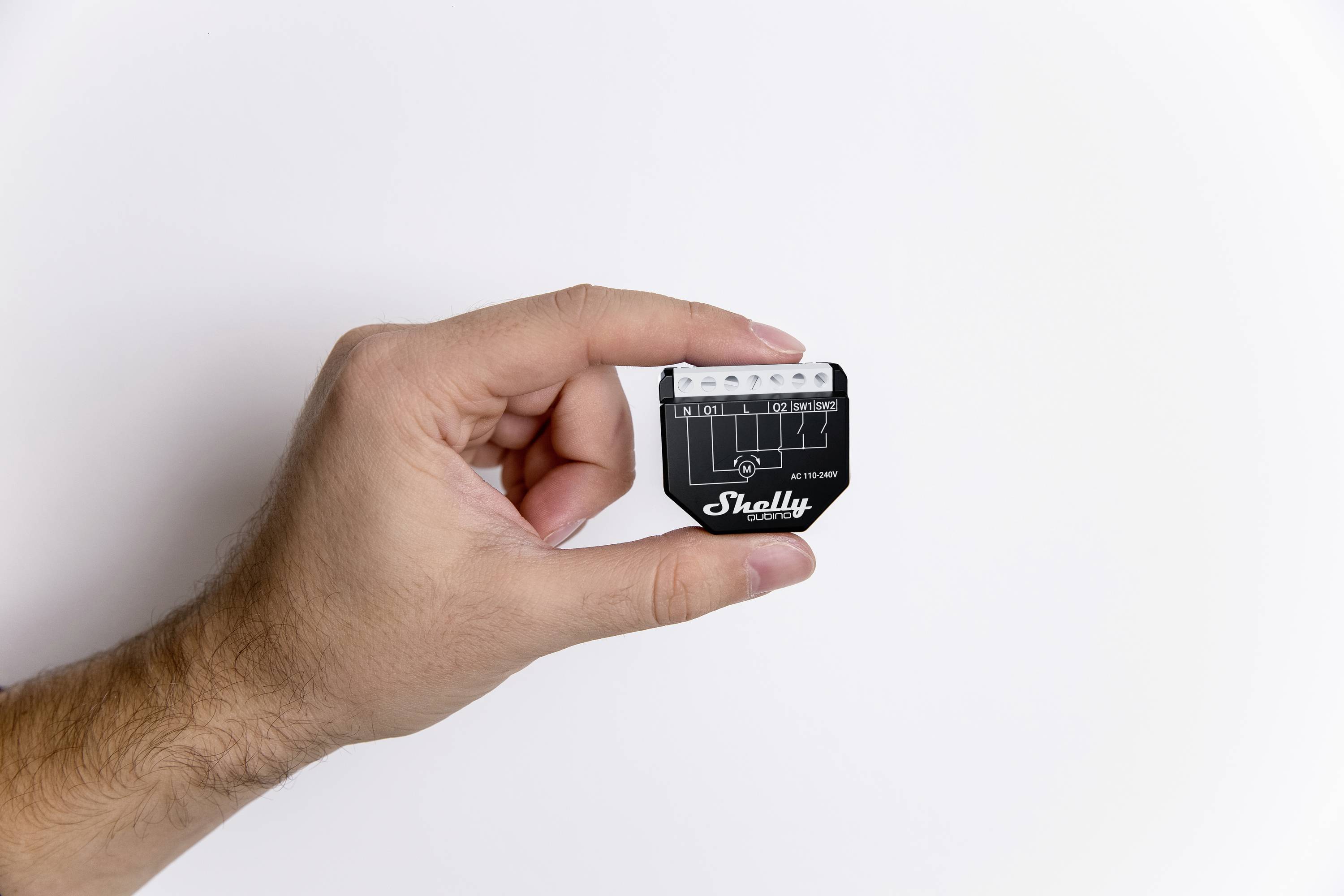 Een hand houdt een klein, zwart elektronisch apparaat vast met het opschrift 'Shelly'. Het apparaat heeft verschillende aansluitingen bovenaan.