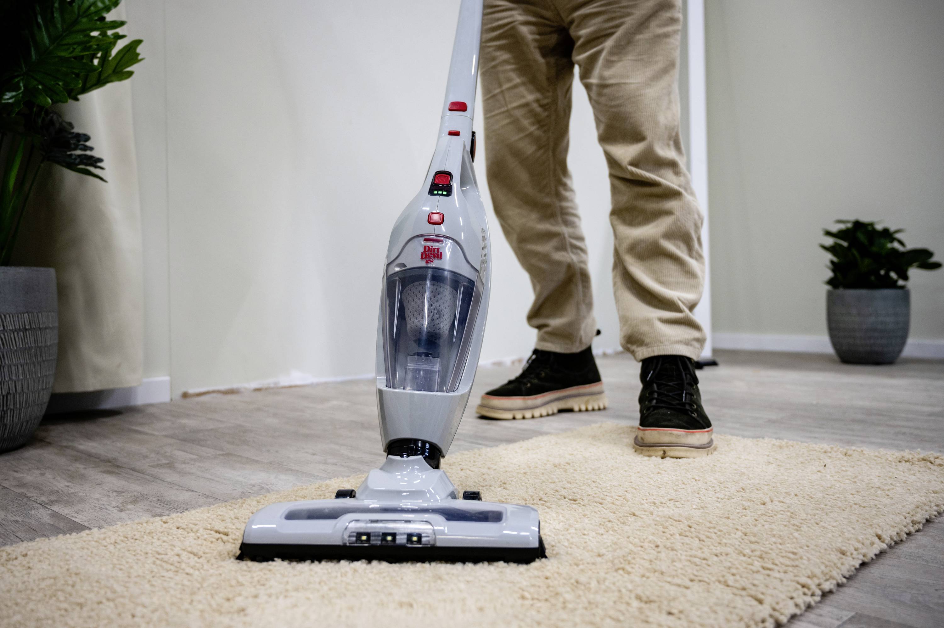 Een persoon bedient een stofzuiger op een beige tapijt in een kamer met houten vloer en planten op de achtergrond.