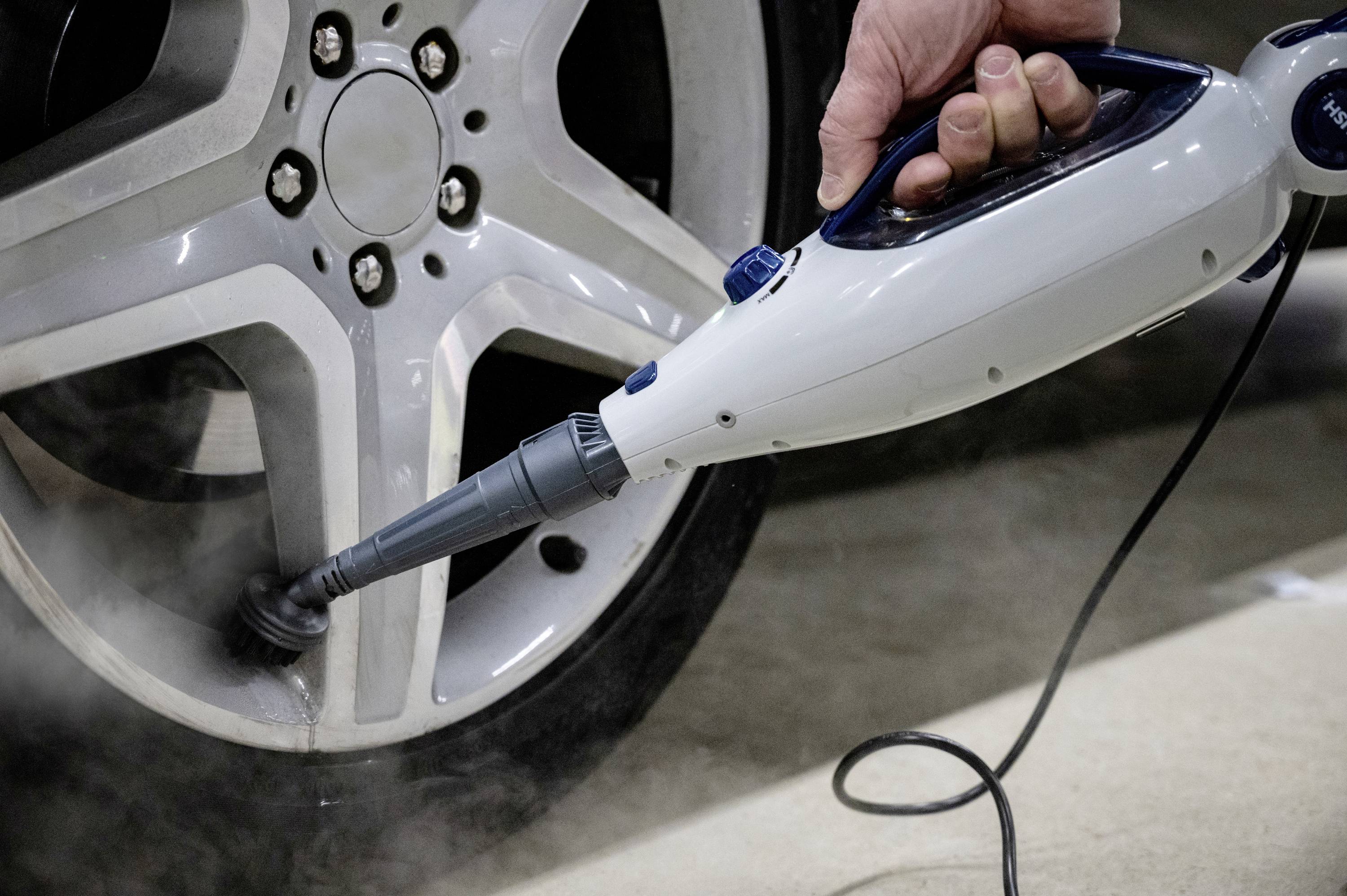 Een persoon reinigt een autovelg met een stoomreiniger. De stoom verwijdert vuil van de wielbouten en de velg.