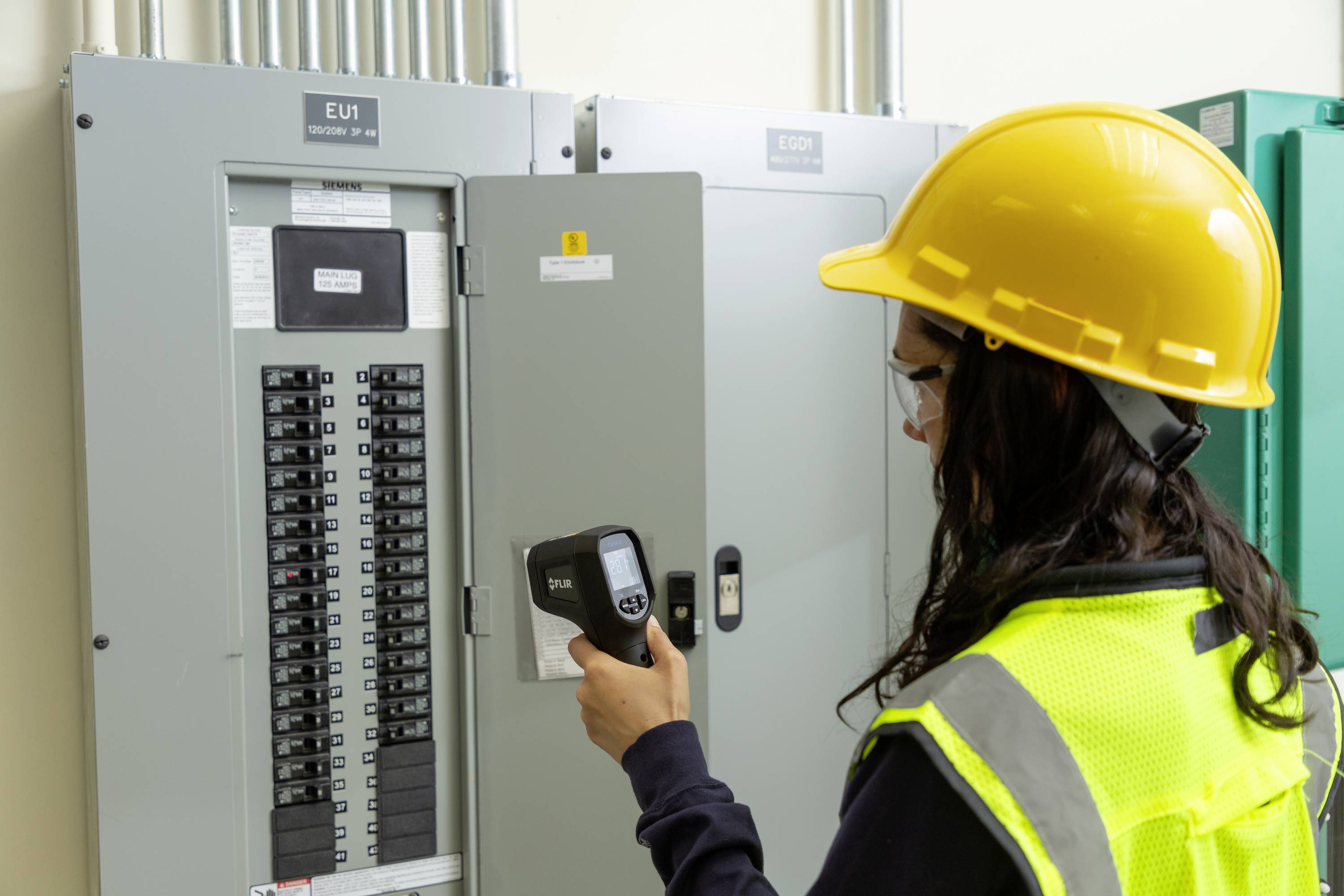 Een persoon met een veiligheidshelm en veiligheidsvest controleert een zekeringenkast in een gebouw met een elektrische meetinstrument.