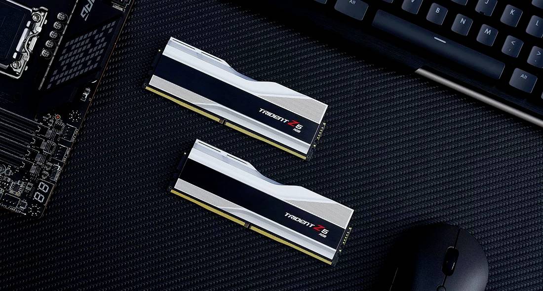 Twee RAM-modules naast elkaar op een zwart oppervlak, naast een toetsenbord en computercomponenten.