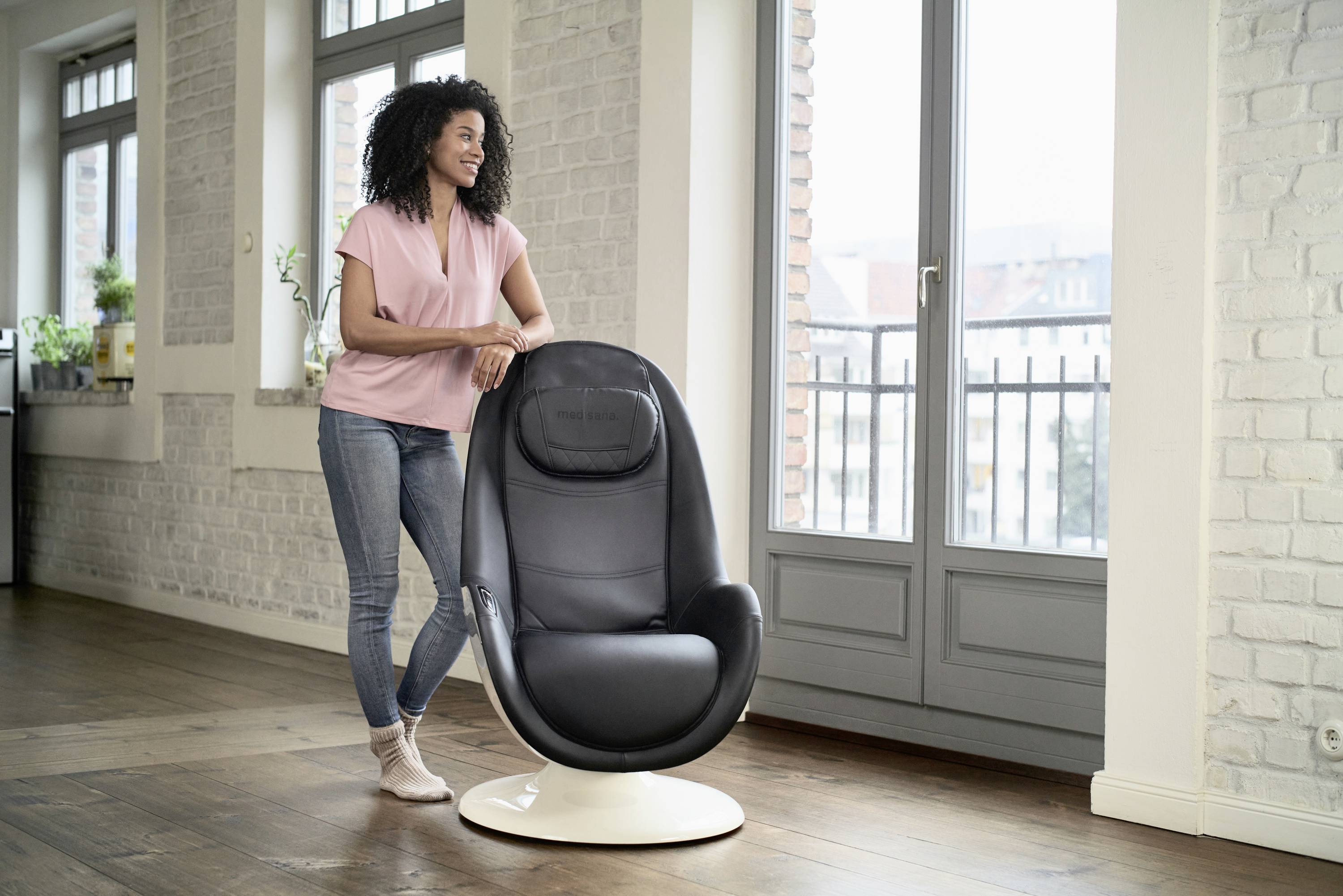 Vrouw leunt glimlachend tegen een zwarte massagestoel in een moderne, lichte ruimte met grote ramen.