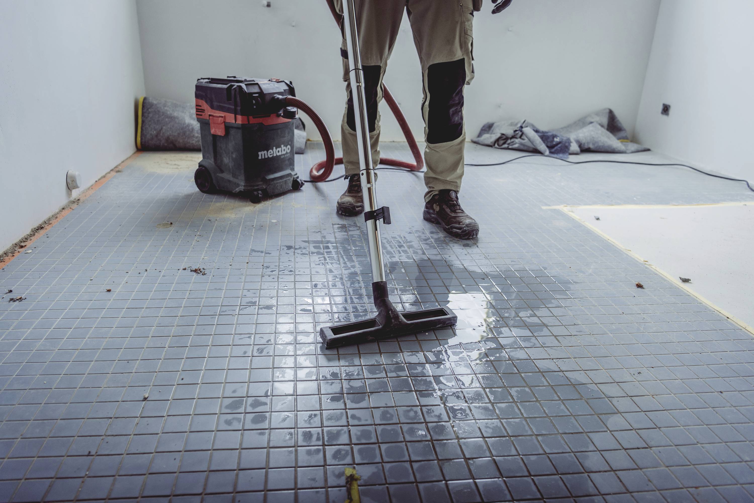 Een persoon reinigt een natte tegelvloer met een stofzuiger in een onafgewerkte ruimte. Bouwmaterialen zijn op de achtergrond zichtbaar.