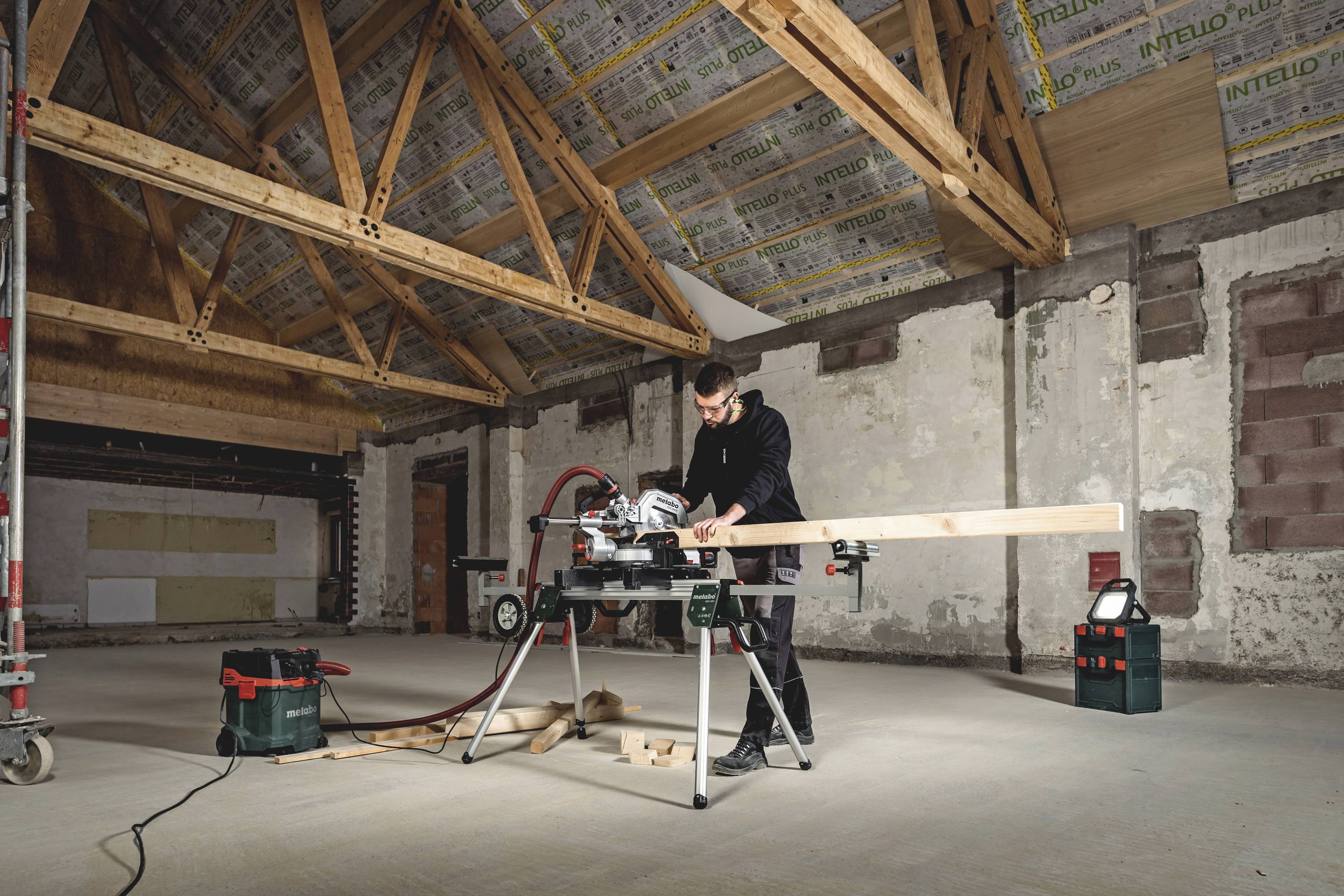 Een persoon werkt aan een tafelzaag in een op het eerste gezicht leeg ogend vertrek, waarbij hij een houten balk doorsnijdt. Een houten balkenzoldering is zichtbaar.