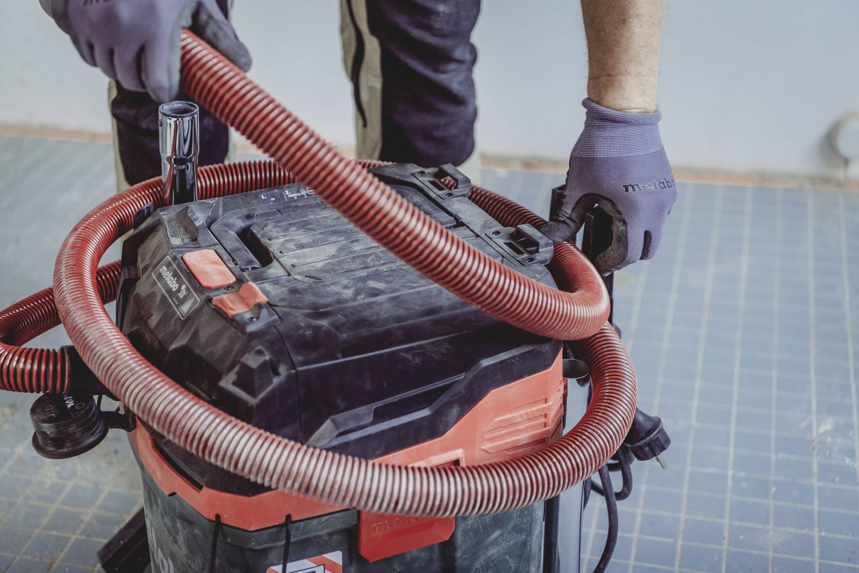 Een persoon sluit een rode slang aan op een industriële stofzuiger. De persoon draagt grijze handschoenen en werkt op een betegelde vloer.
