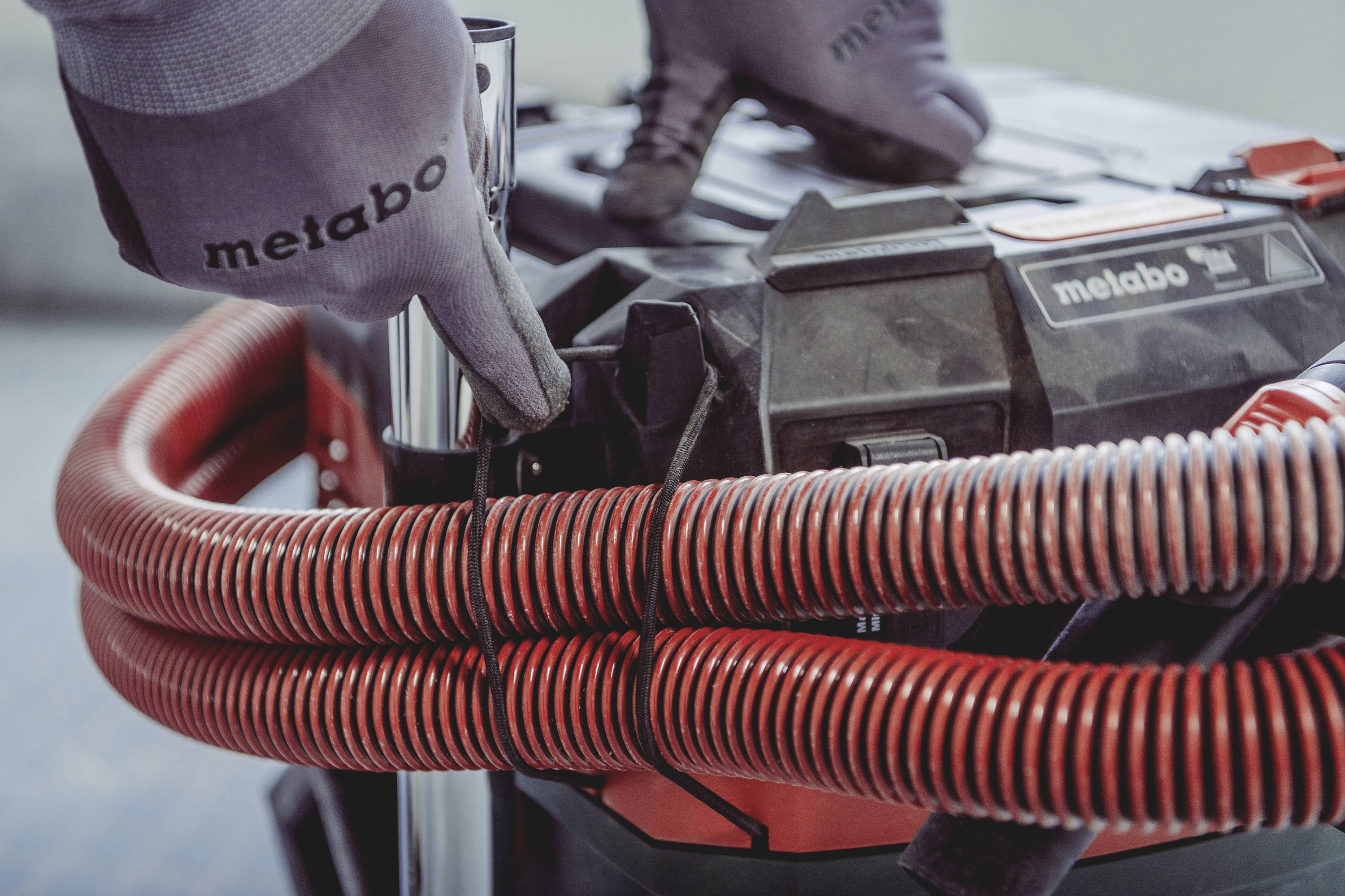 Handschoenen dragen het 'Metabo'-merk, bevestigen rode slang aan zwarte gereedschapskist.