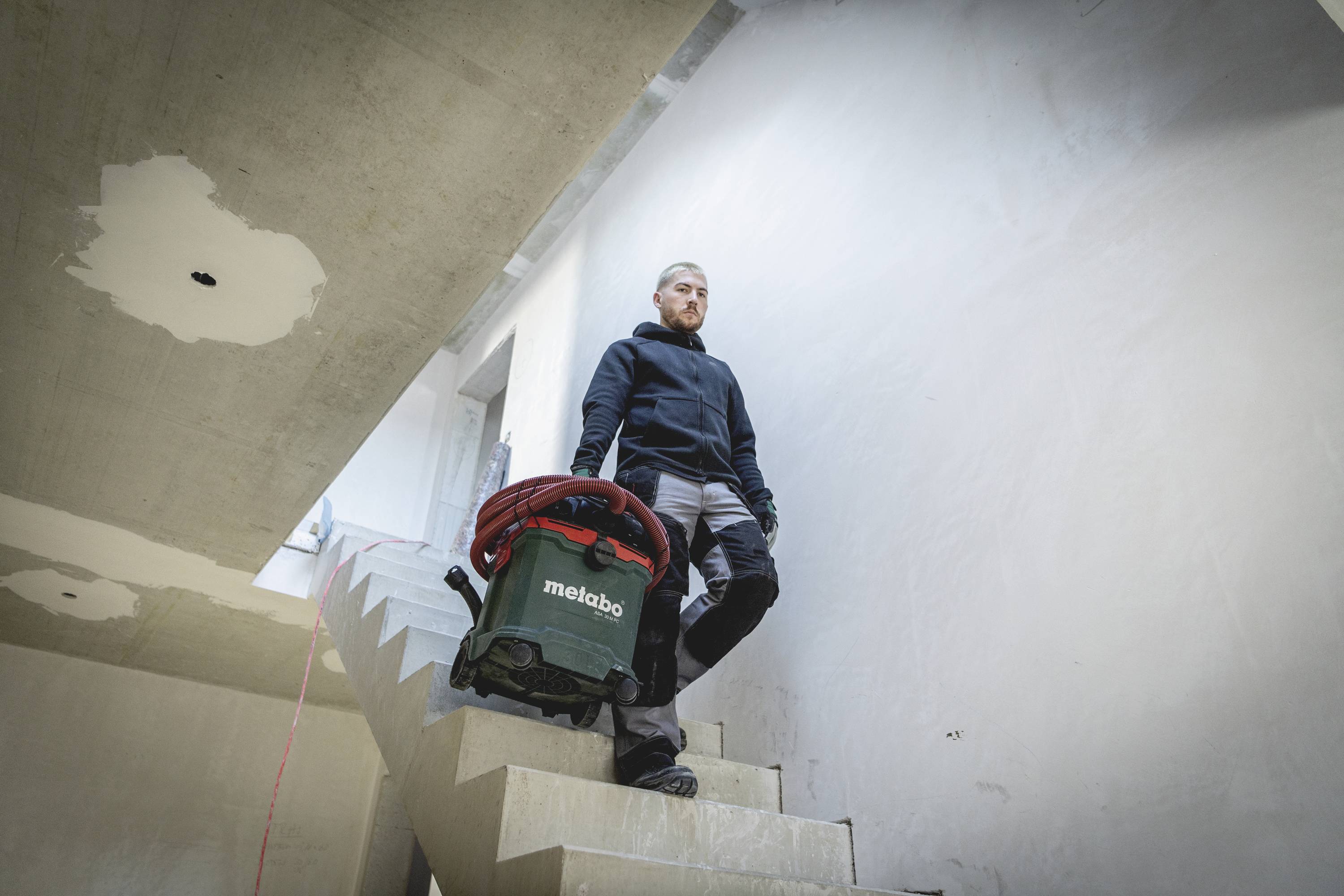Een vakman draagt een stofzuiger een trap af in een onafgewerkt gebouw. De omgeving laat onbepleisterde muren zien.