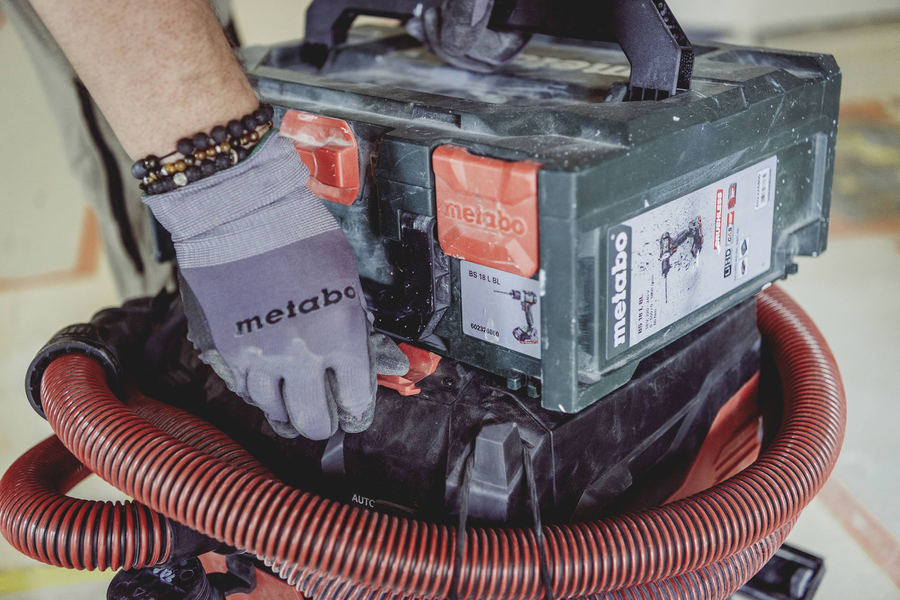 Een ambachtsman draagt handschoenen en bedient een stofzuiger-achtig apparaat van Metabo. Een rode slang is verbonden met het apparaat.
