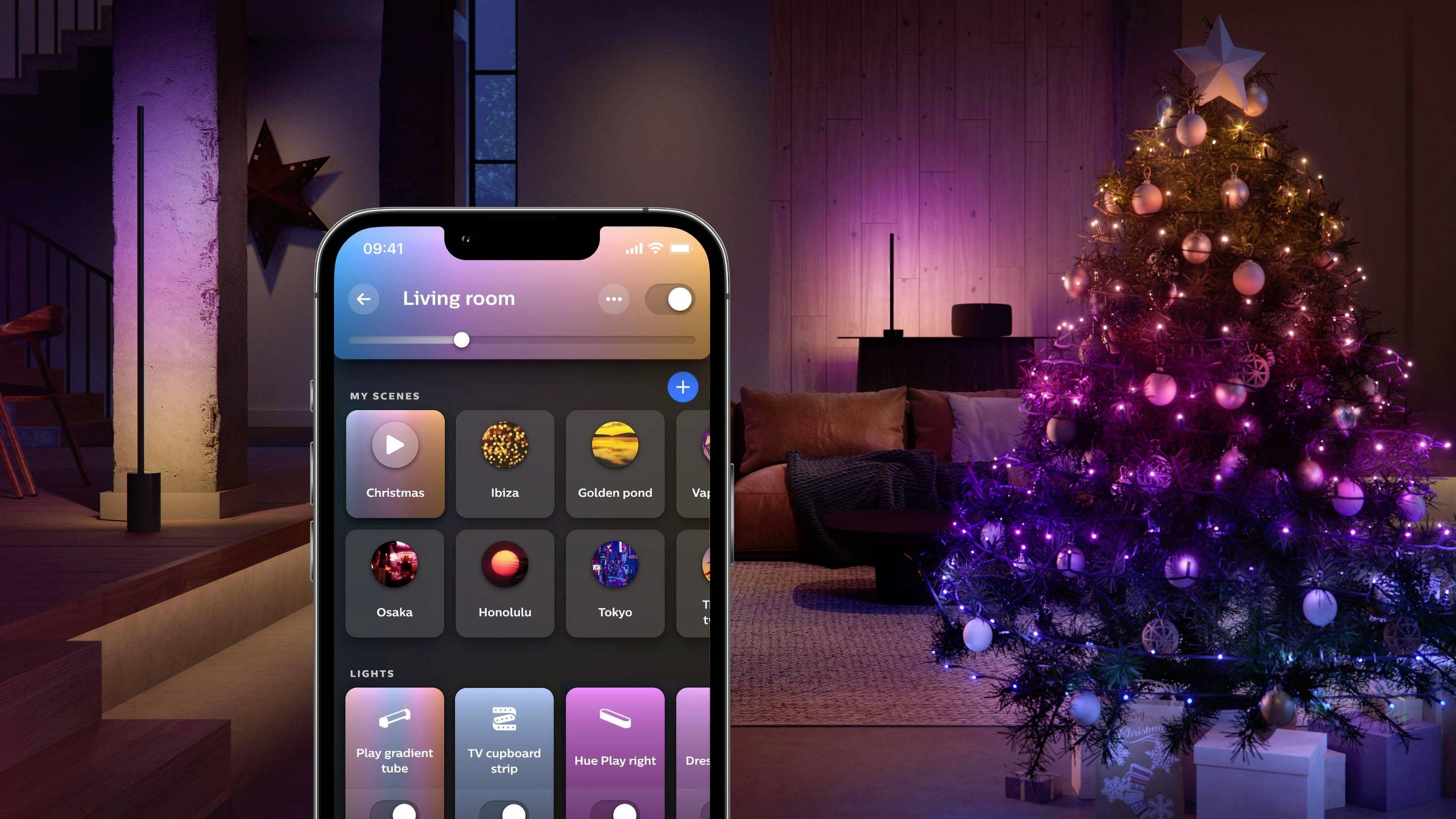 Een smartphone toont een app voor het besturen van lichtsferen in de woonkamer. Rechts staat een kerstboom met kleurrijke verlichting.