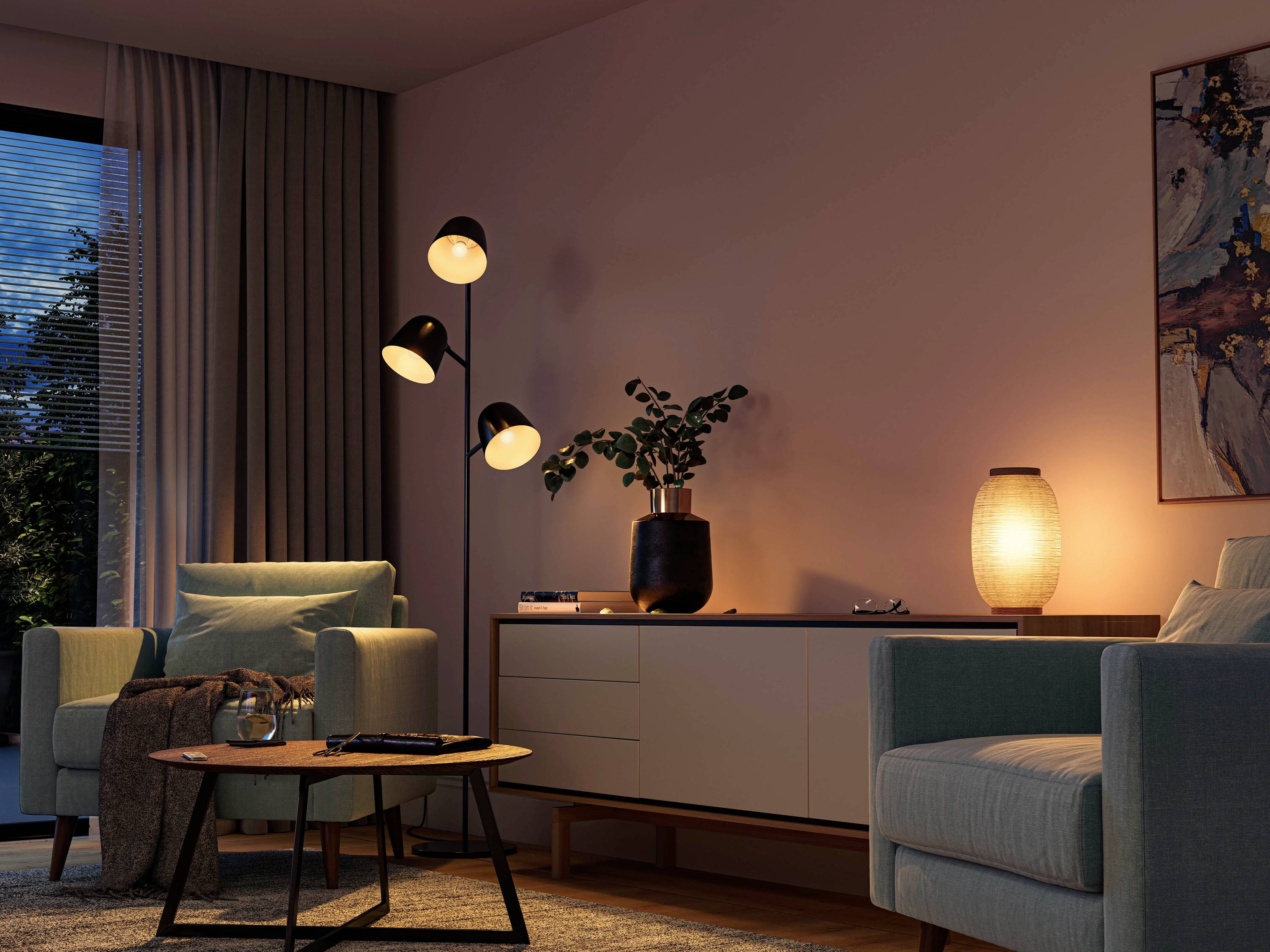 Gezellige woonkamer bij schemering, twee fauteuils, ronde tafel, staande lamp, dressoir met plant en lamp, groot raam links.