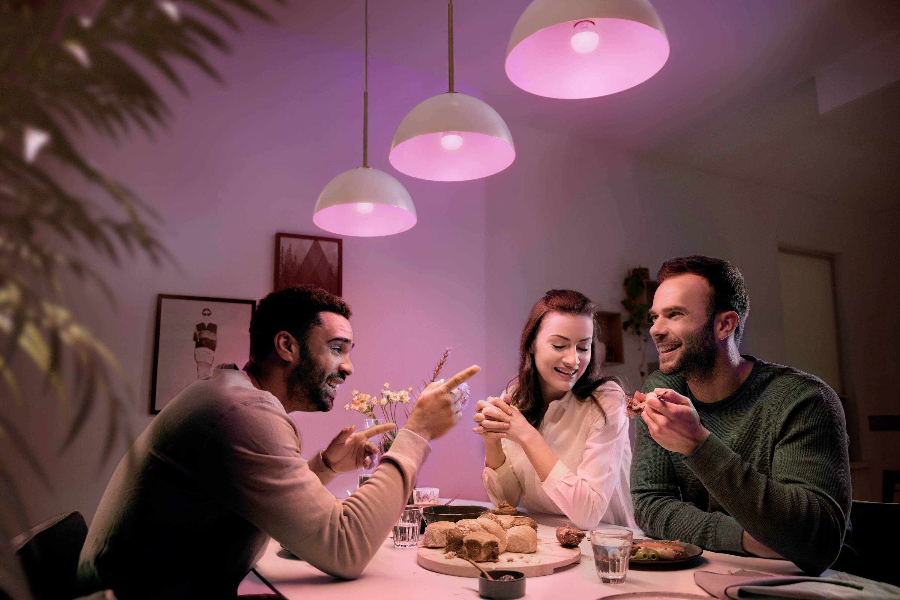 Drie personen zitten aan een tafel met eten, lachen en praten met elkaar. De ruimte is warm verlicht, met hangende lampen.