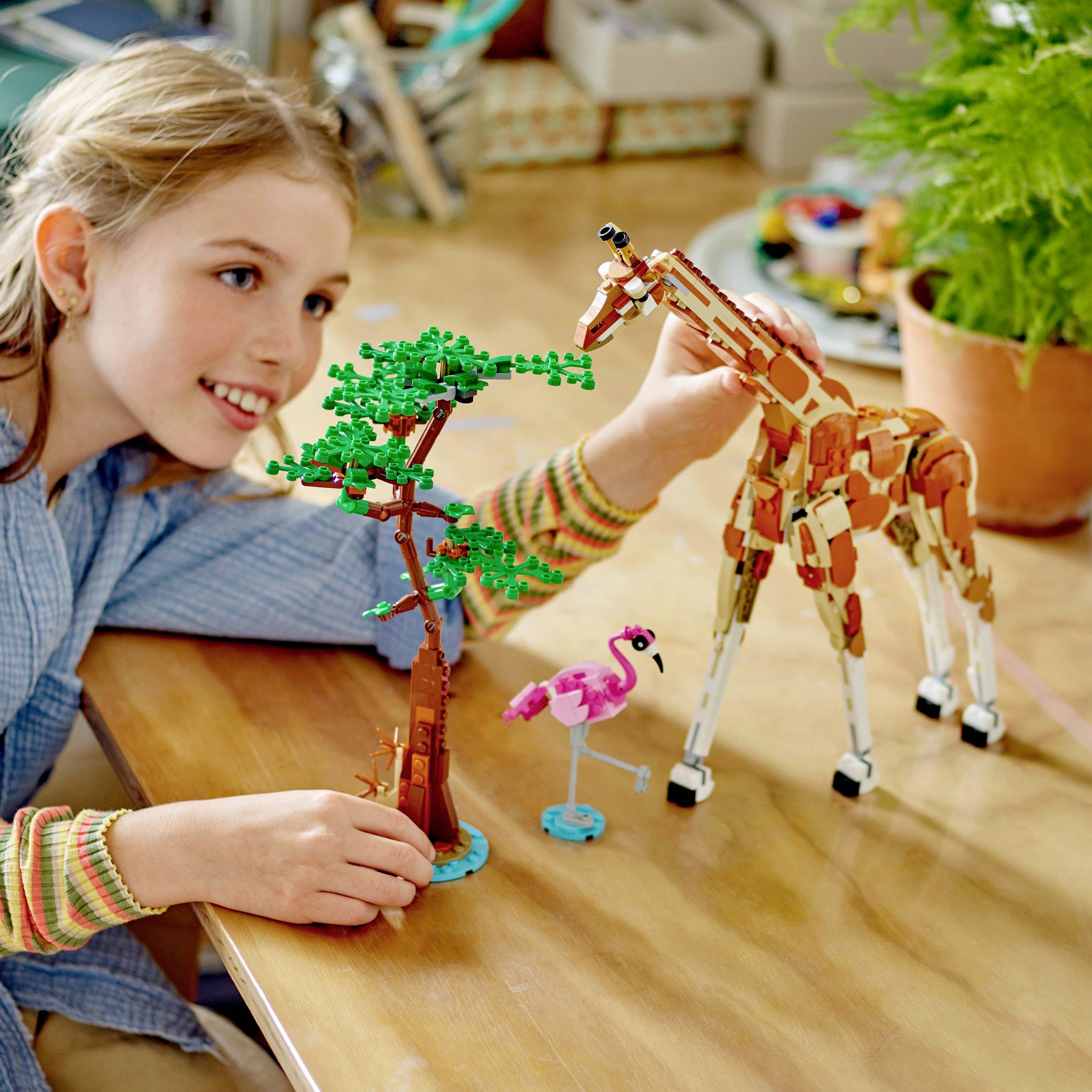 Een kind speelt met Lego-dierfiguren, waaronder een giraffe en een boom. Op de achtergrond zijn meer speelgoed en een plant te zien.