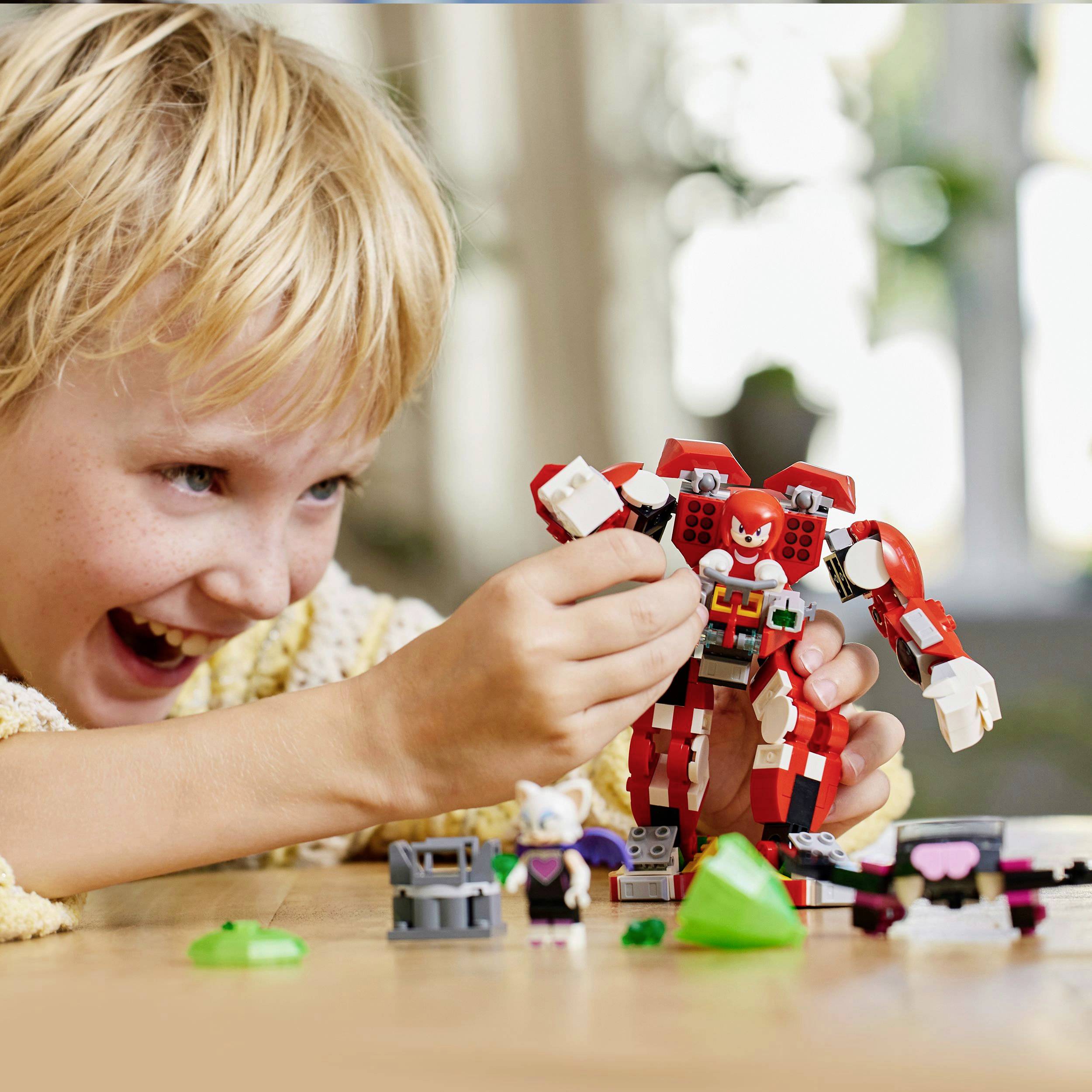Een lachend kind speelt met een rood-wit robot-speelgoed op een houten tafel. Op de achtergrond wazig groene planten.