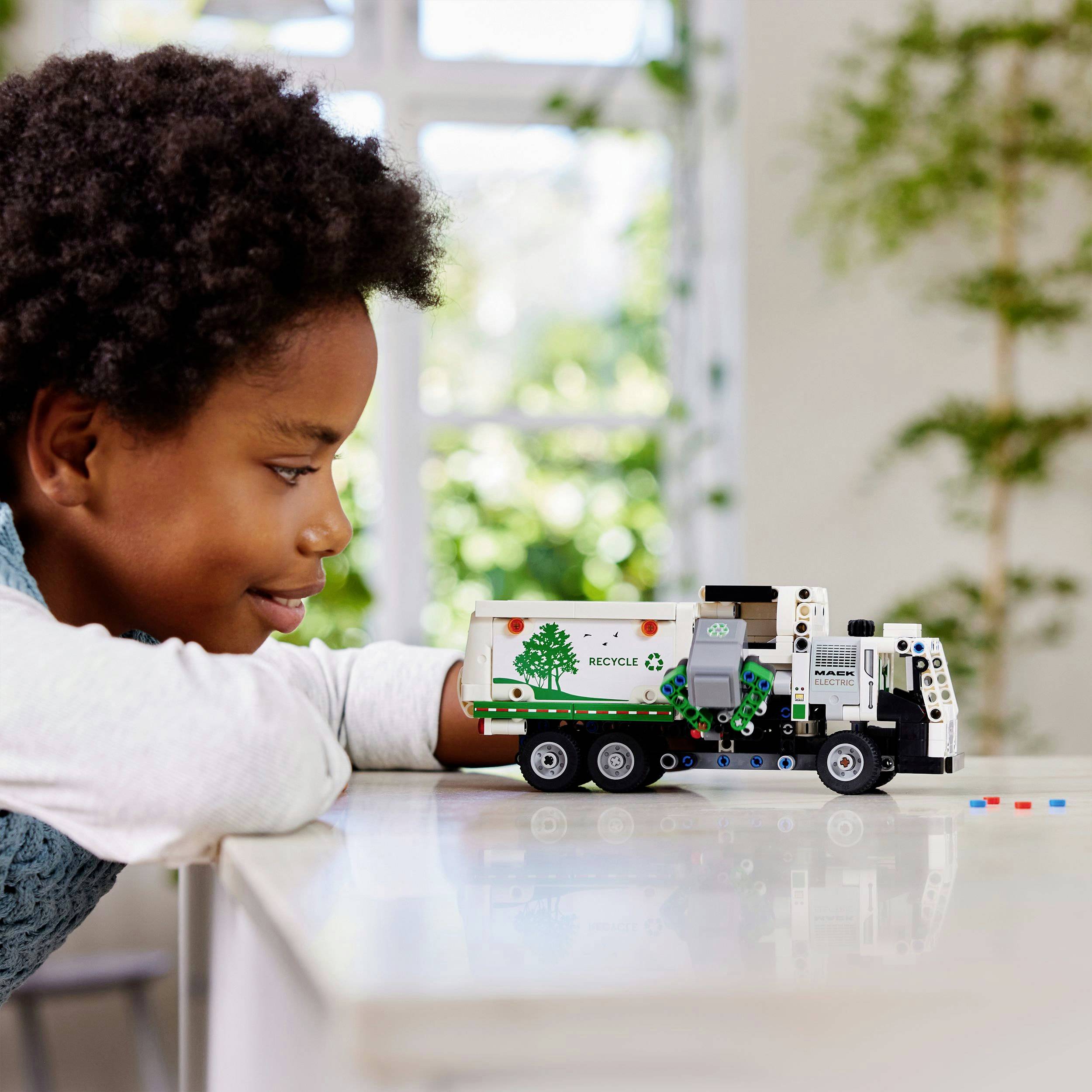 Een kind speelt met een kleine model-vrachtwagen met het thema recycling op een tafel. Op de achtergrond zijn groene planten te zien.