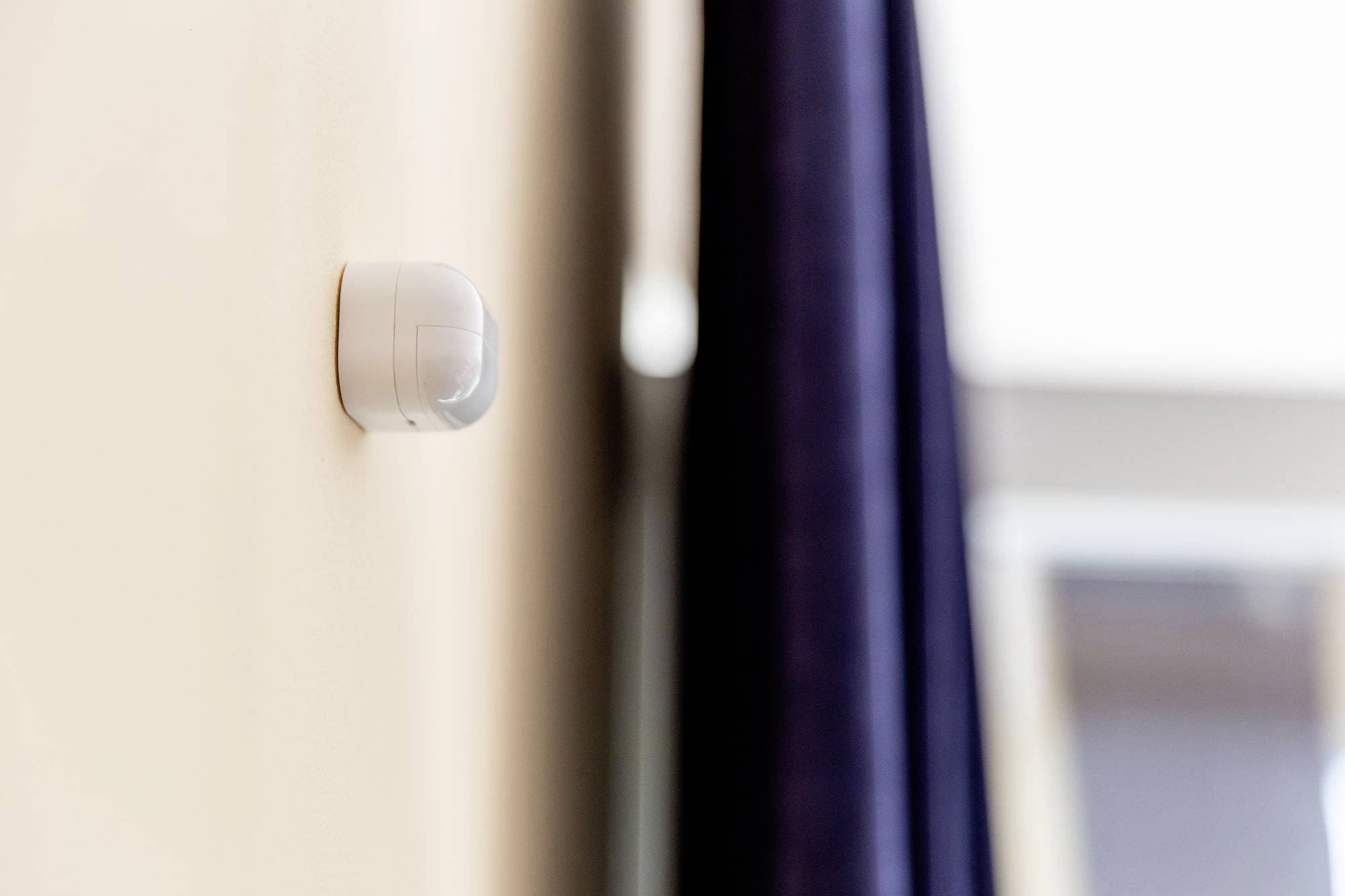 Aan een muur van de kamer is een bewegingsdetector gemonteerd. Op de achtergrond is een raam zichtbaar, dat gedeeltelijk wordt bedekt door een gordijn.