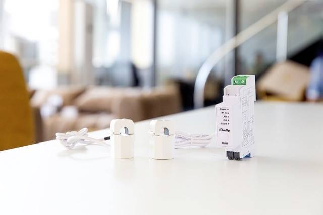 Een energiemeter met kabels ligt op een tafel in een modern kantoor. Op de achtergrond is vaag een bank te zien.