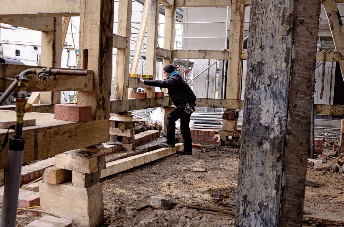 Een bouwvakker in beschermende werkkleding werkt aan een houten steiger op een bouwterrein, omgeven door houten planken en bouwmachines.