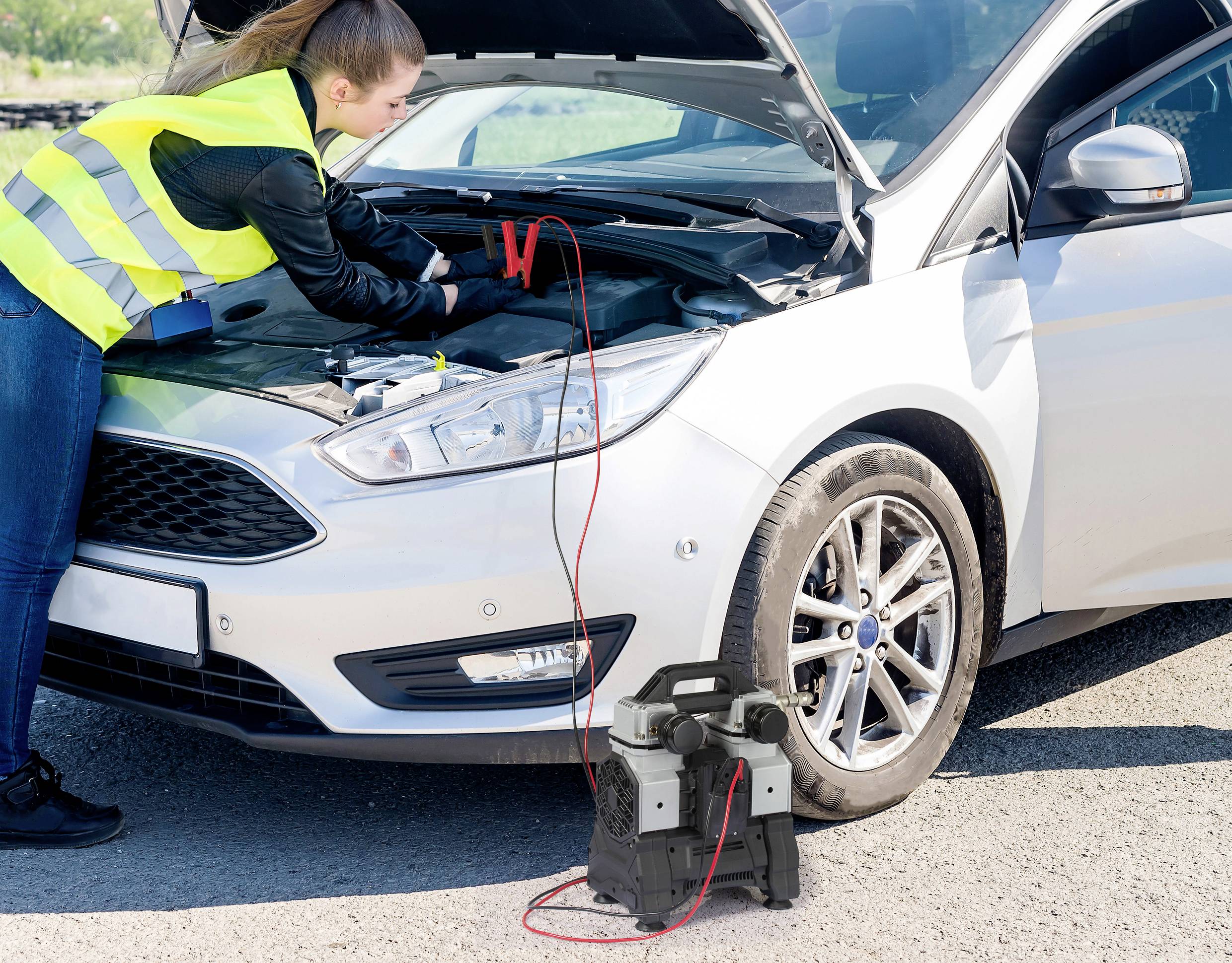 Een vrouw in een gele veiligheidsvest start de auto met een startkabel op een weg. De motorkap staat open.