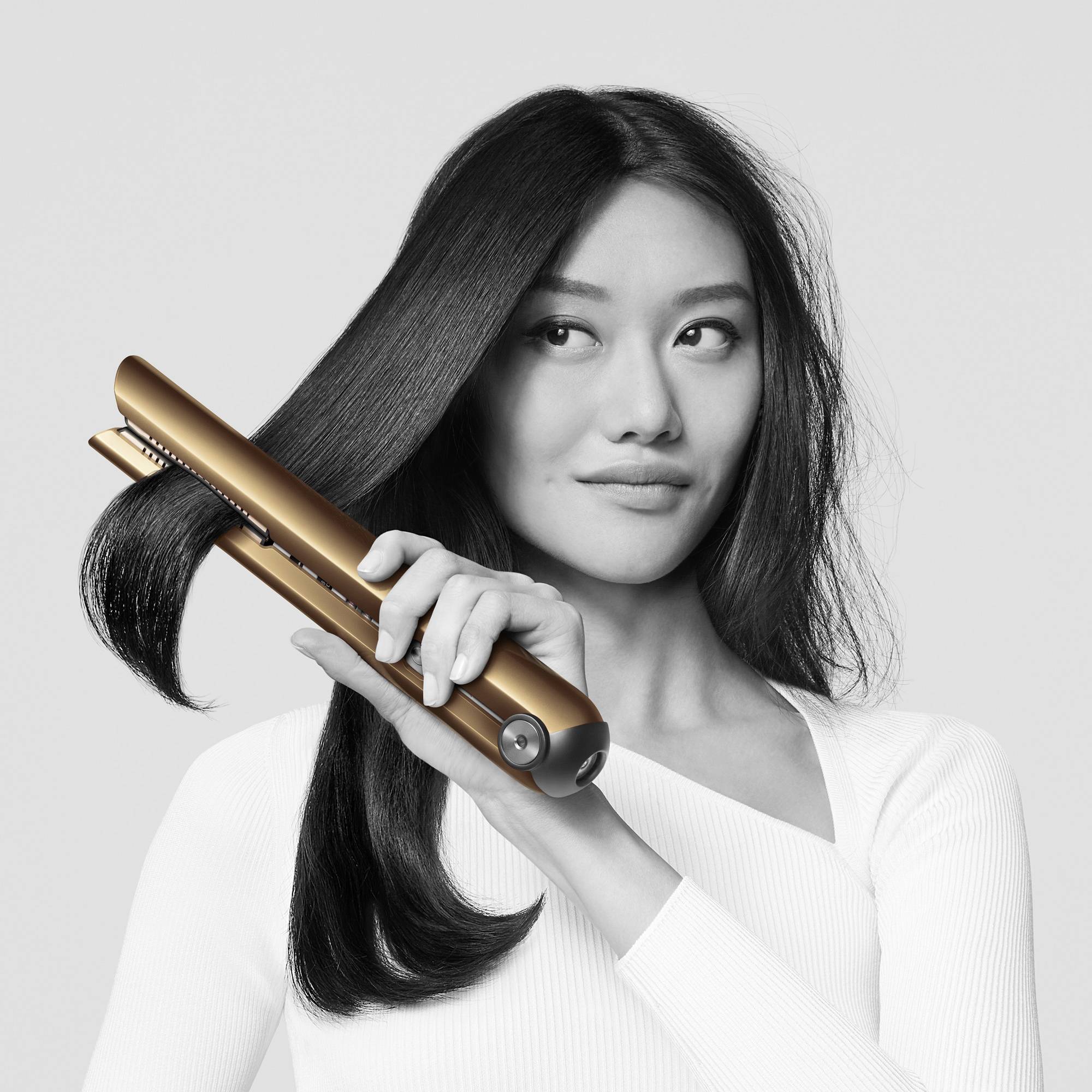 Een vrouw met lang haar strijkt haar haar glad met een gouden stijltang. Ze draagt een wit bovenste kledingstuk en kijkt licht opzij.