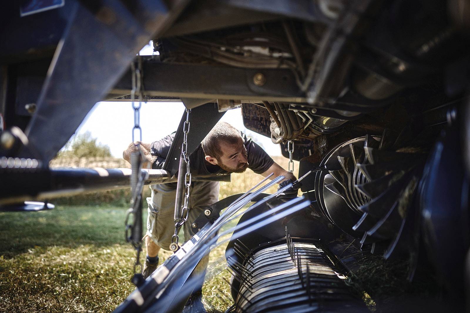 Een man werkt onder een landbouwmachine. Hij controleert de mechaniek ervan. De omgeving is een weide op een zonnige dag.